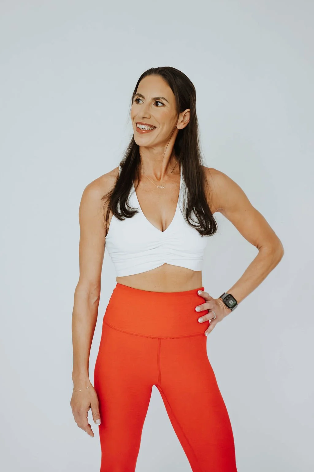 A woman with long dark hair wearing a white sports crop top, red workout leggings, and a fitness tracker on her wrist, standing against a plain light background.