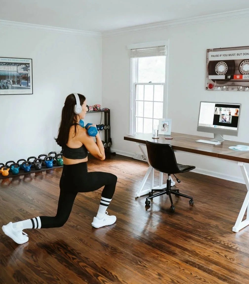 A woman is working out at home, holding a dumbbell in each hand, with a desk and computer in the background.