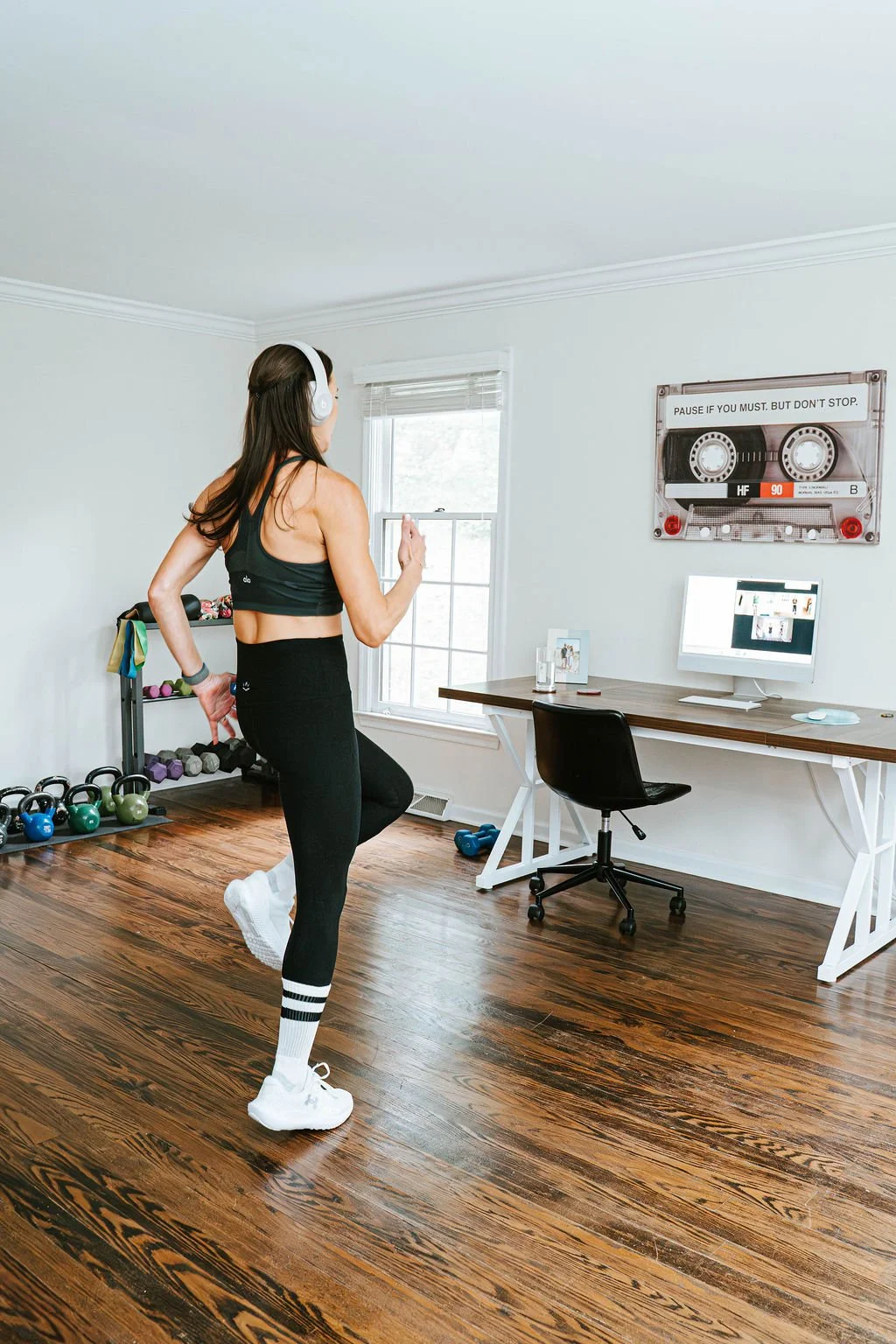 A woman exercising in a home gym, wearing workout clothes and headphones, with dumbbells and kettlebells on a rack behind her, and a computer on a desk on the right side of the room.