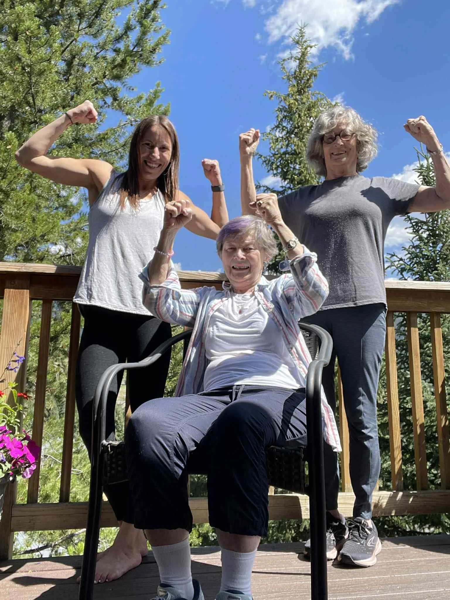 Three women outdoors on a sunny day, flexing their muscles and smiling. One woman is sitting in a chair, and two women are standing behind her, raising their fists in a sign of strength. There are trees and a blue sky in the background.