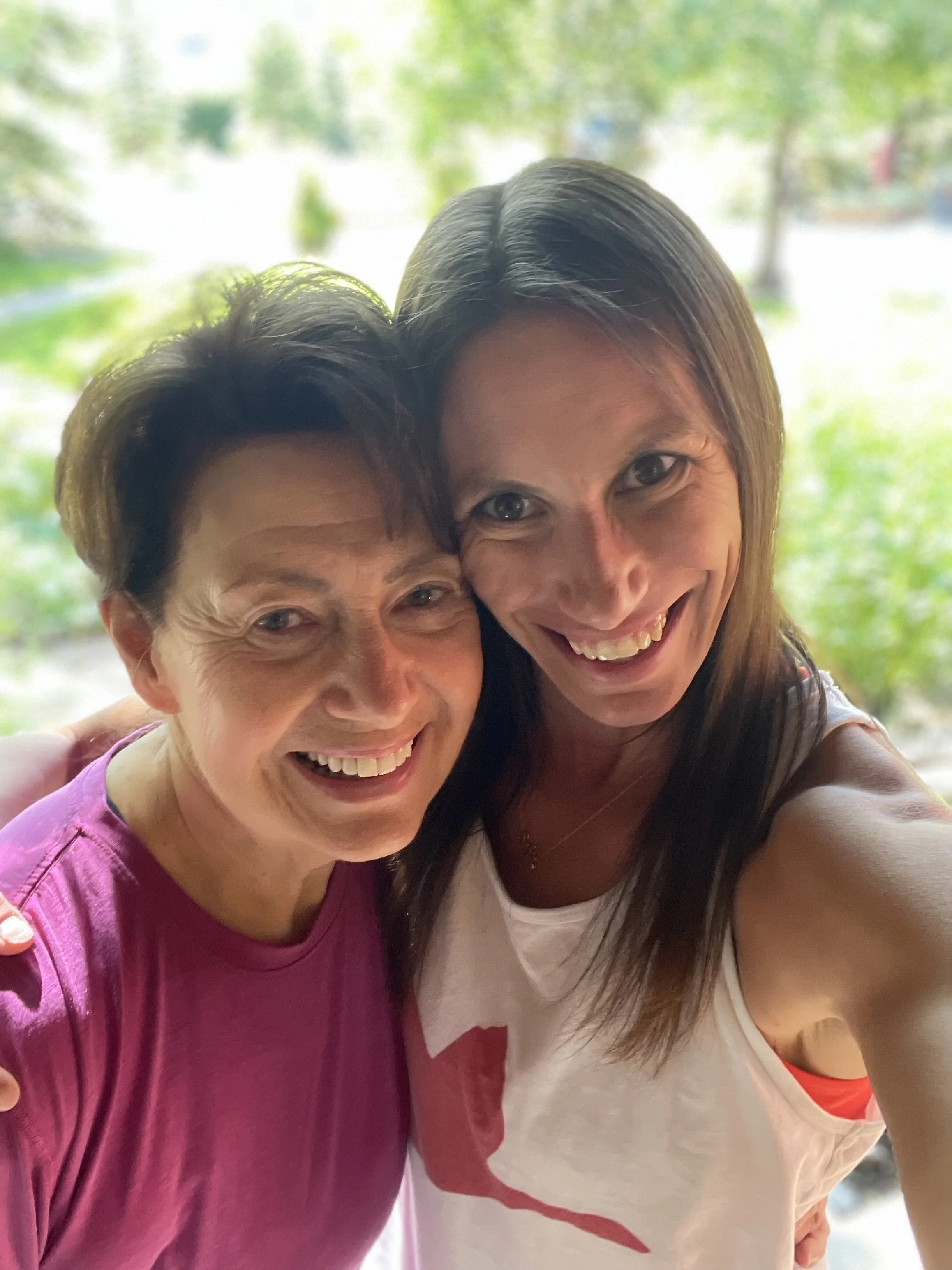 Two women taking a selfie outdoors, smiling, with greenery in the background.