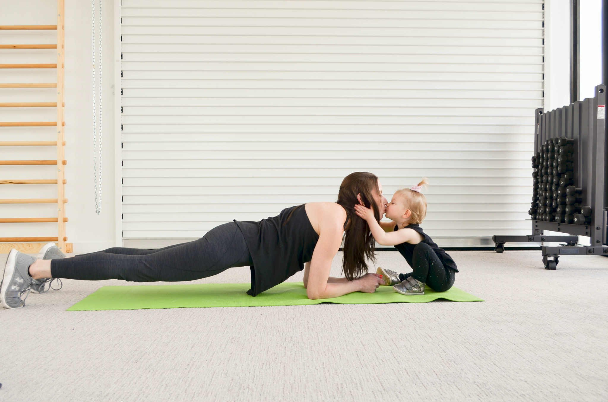 A woman doing a plank exercise on a green yoga mat while a young girl hugs and kisses her in a gym or fitness studio.