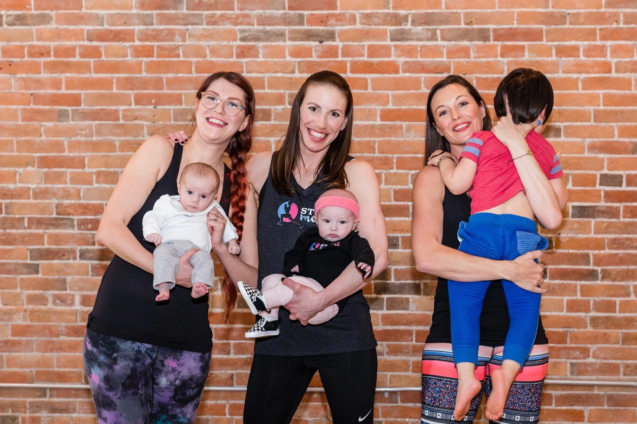 Four women smiling with three babies against a brick wall, some women holding babies and one woman hugging a child.