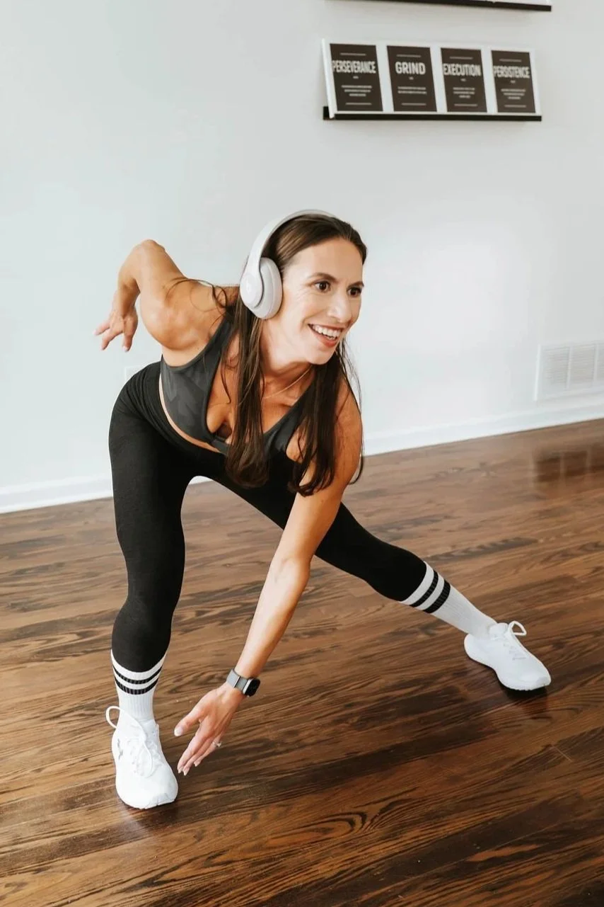 A woman with long brown hair wearing headphones, a black athletic outfit, and white sneakers, smiling while doing a workout stretch on hardwood floor in a fitness studio.