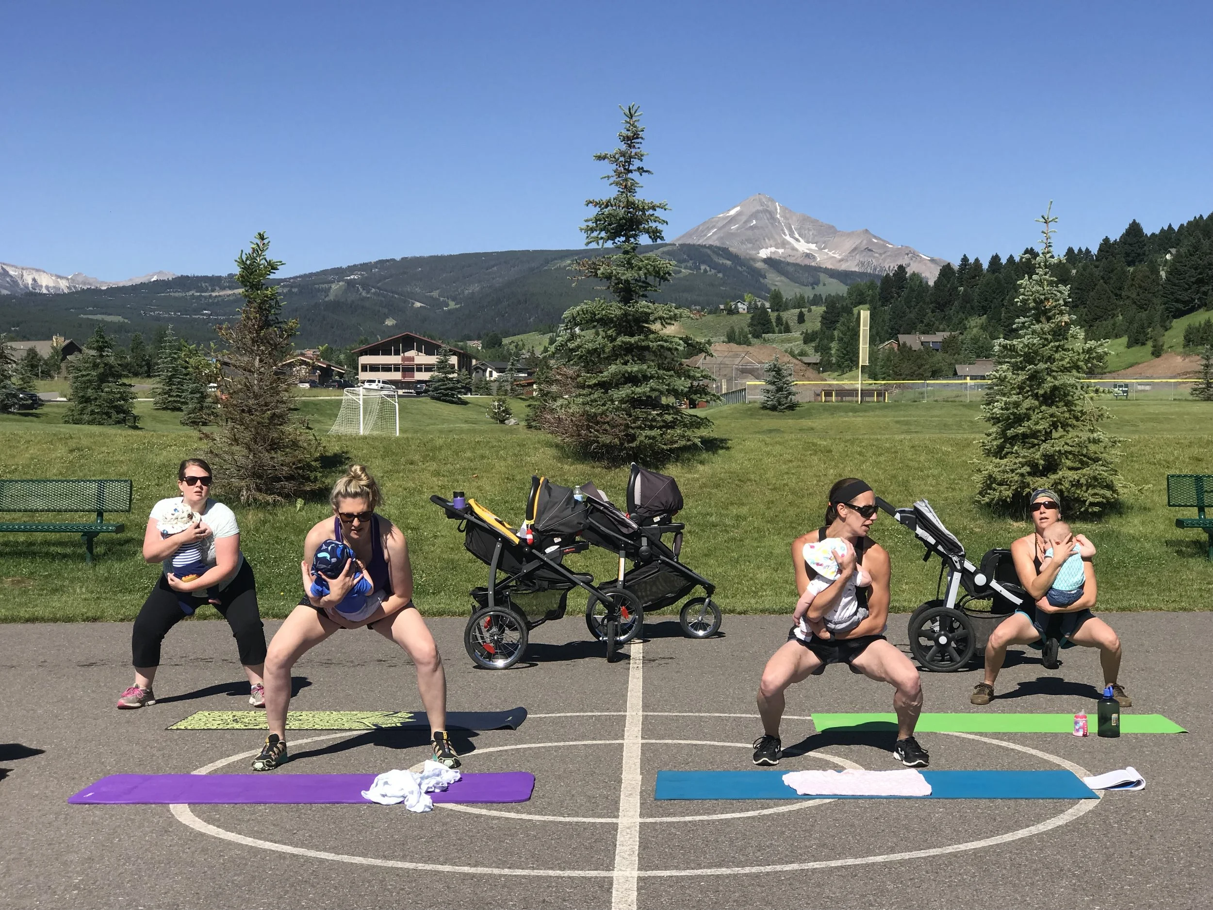 Four women exercising outdoors on yoga mats on a basketball court, holding babies in their arms, with strollers and a scenic mountain view in the background.