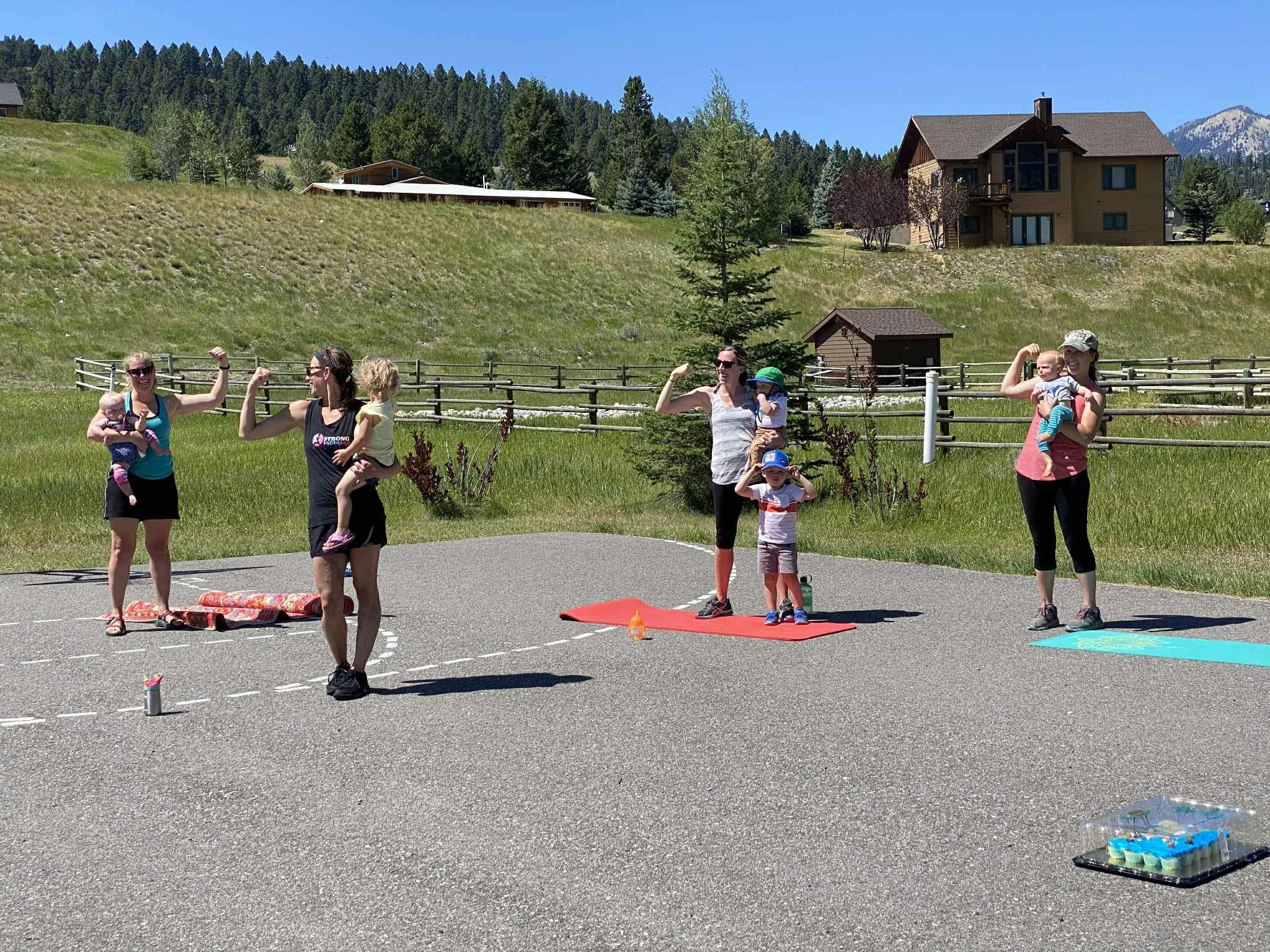 Four women with children participating in outdoor fitness exercises on a paved area with grass, trees, and houses in the background on a sunny day.