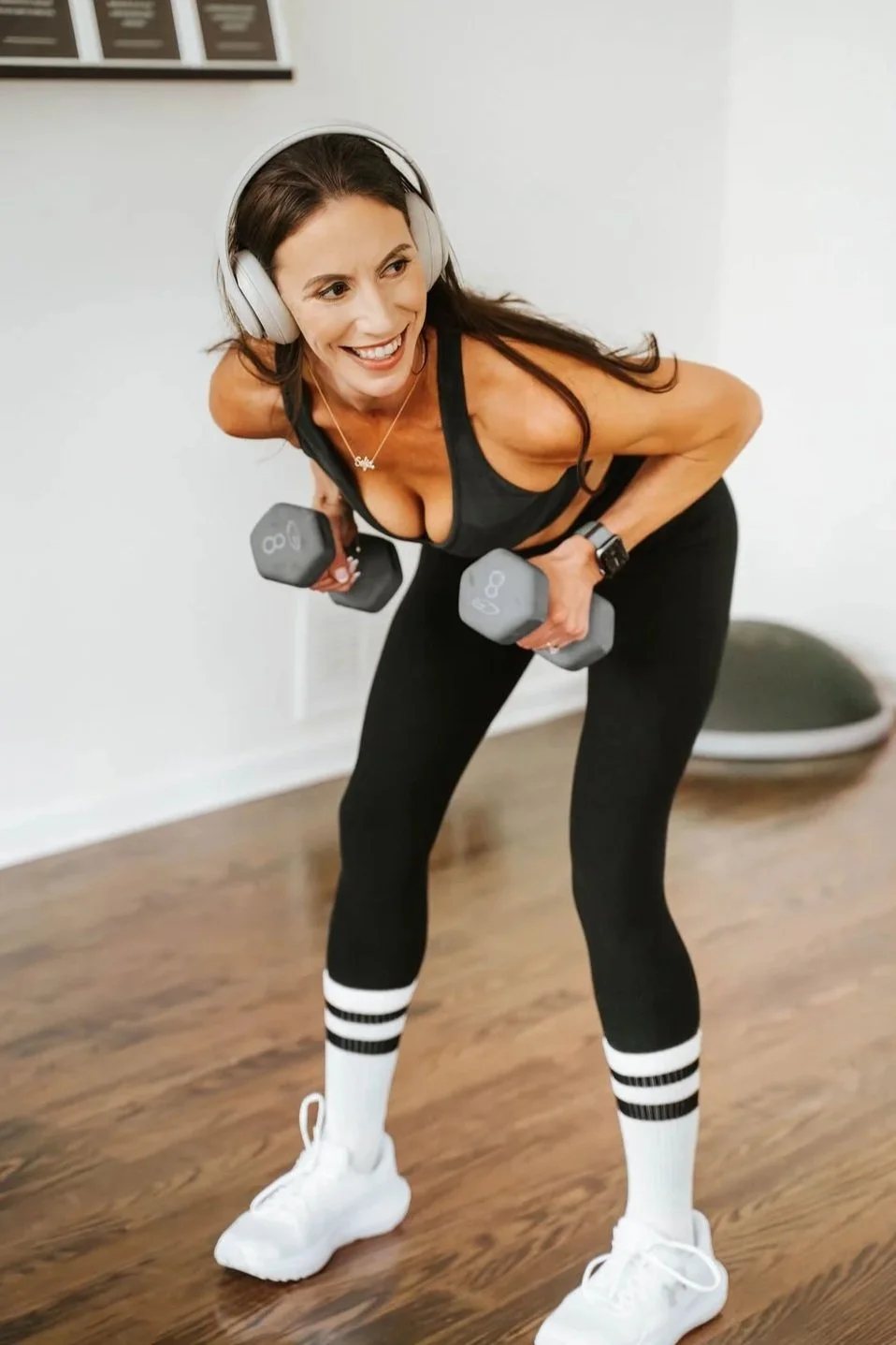 A woman with long dark hair, wearing headphones, a black sports bra, black leggings with white striped socks, and white athletic shoes, is smiling and lifting dumbbells while working out indoors on a wooden floor.