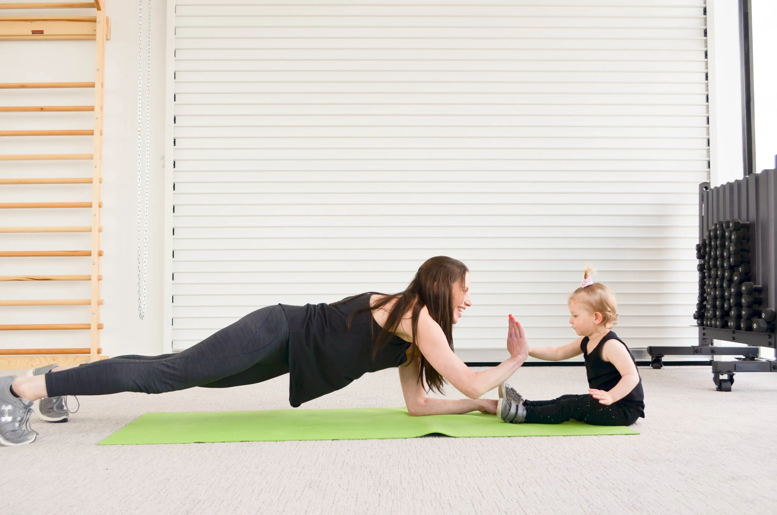 Woman and young girl doing a yoga plank exercise together, giving each other a high five on a green yoga mat in a gym.