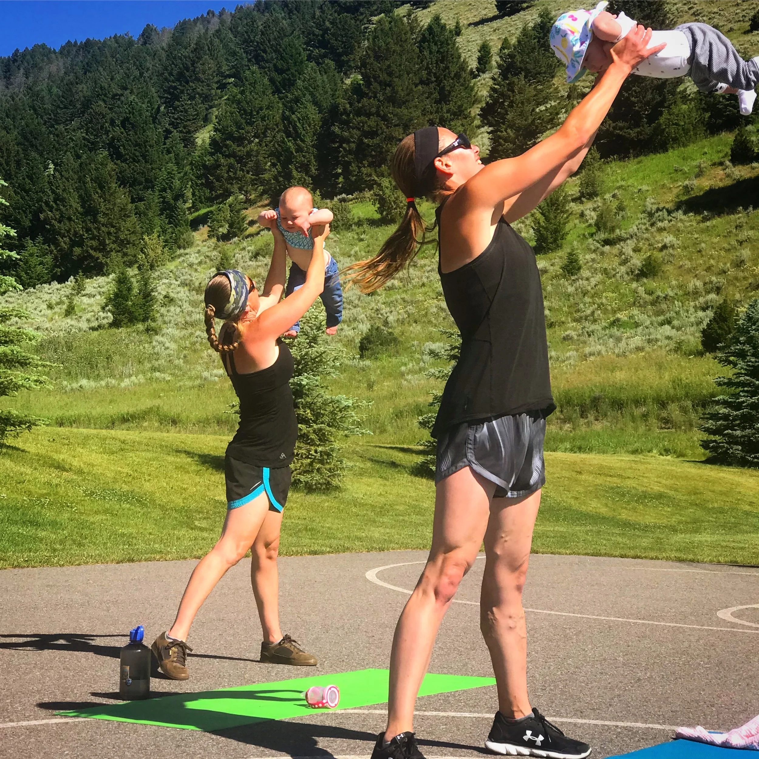 Two women and a child exercising outdoors on a basketball court. One woman is lifting the child, and the other woman is doing a plank while holding a baby doll. The setting is surrounded by green grass, trees, and mountains.