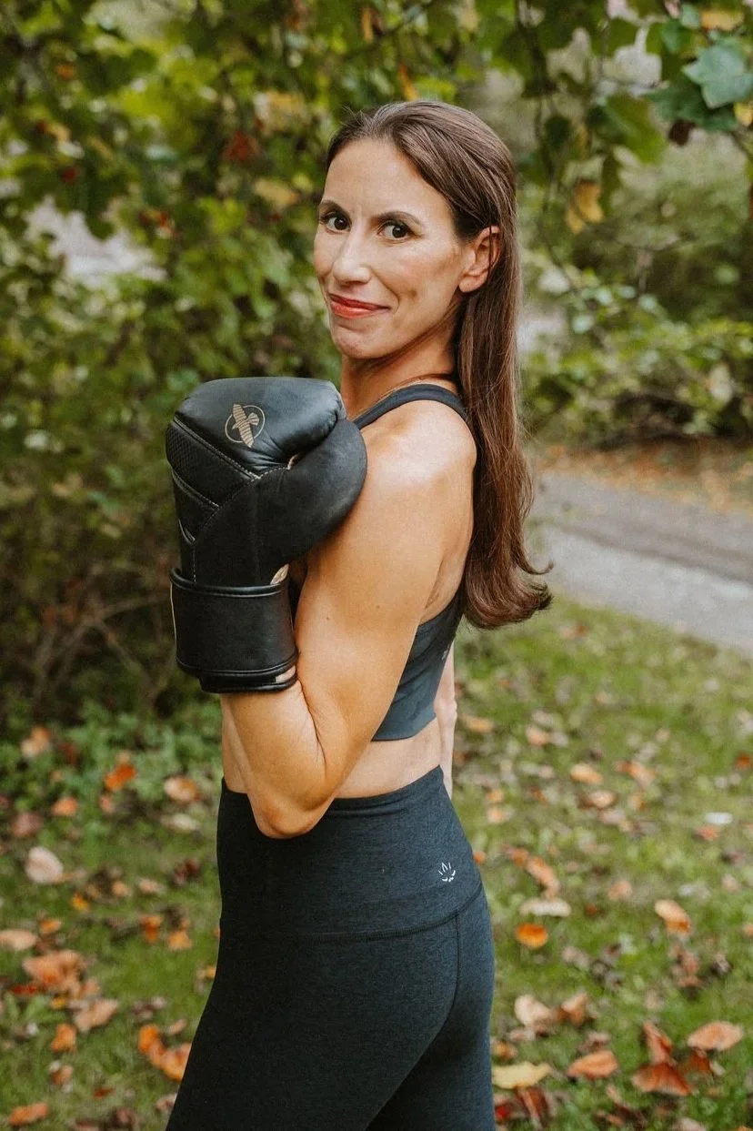 A woman with long brown hair, wearing workout clothes and boxing gloves, stands outdoors in a natural setting with trees and fallen leaves, looking over her shoulder with a small smile.