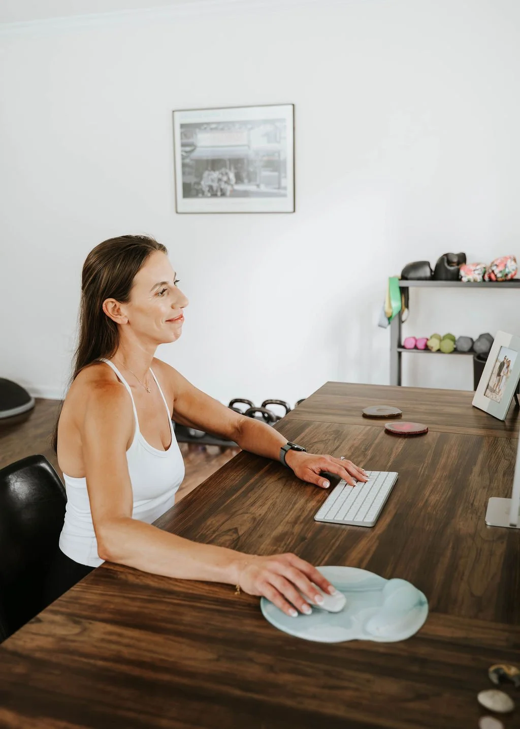 A woman sitting at a wooden desk working on a computer in a home gym or exercise space, with dumbbells and fitness equipment in the background.