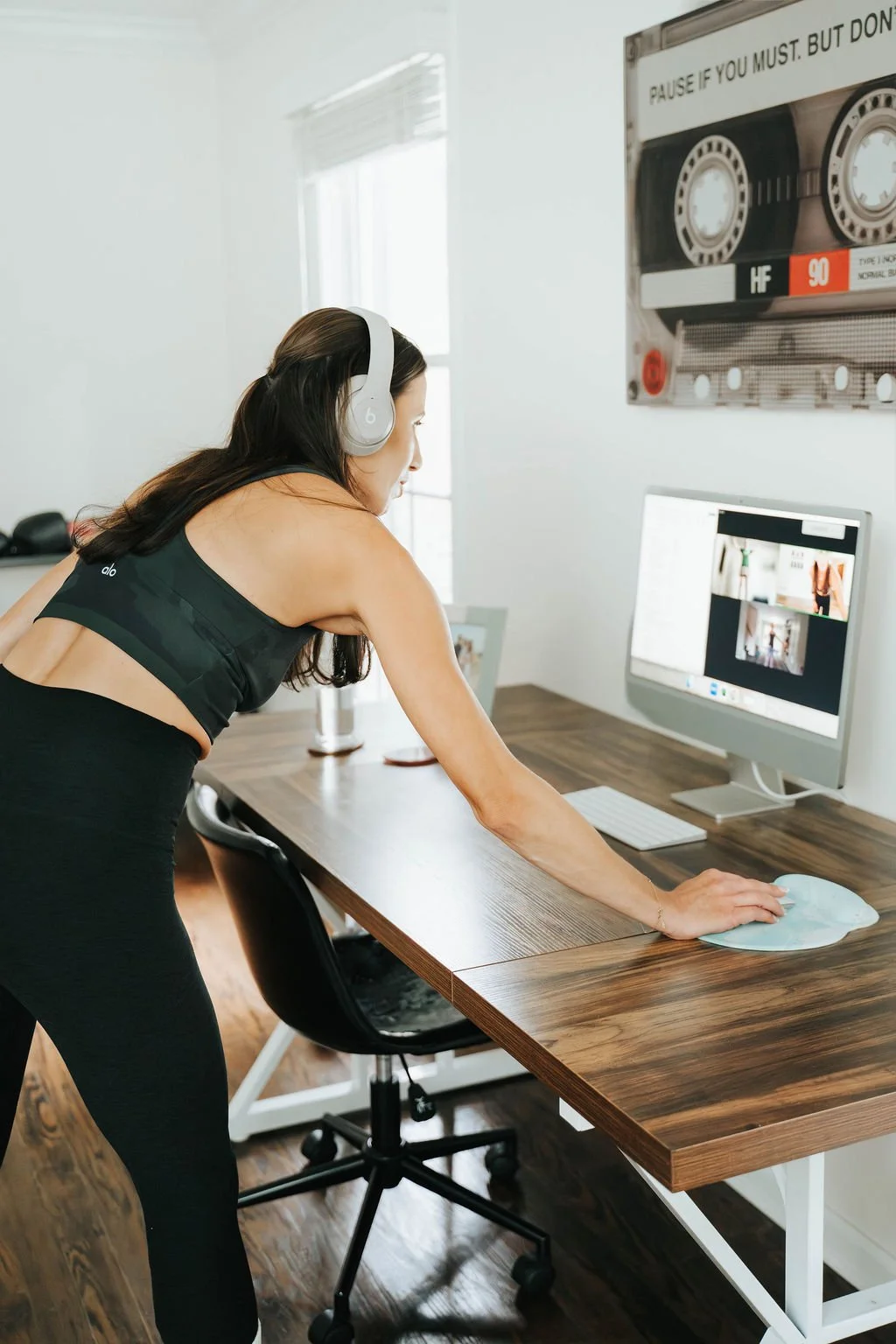 Woman with headphones cleaning a computer monitor at a wooden desk in a bright room.