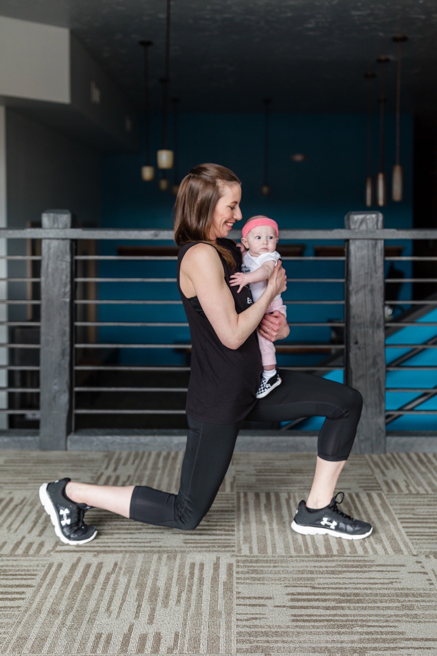 A woman performing a lunge exercise indoors, holding a baby in her arms.