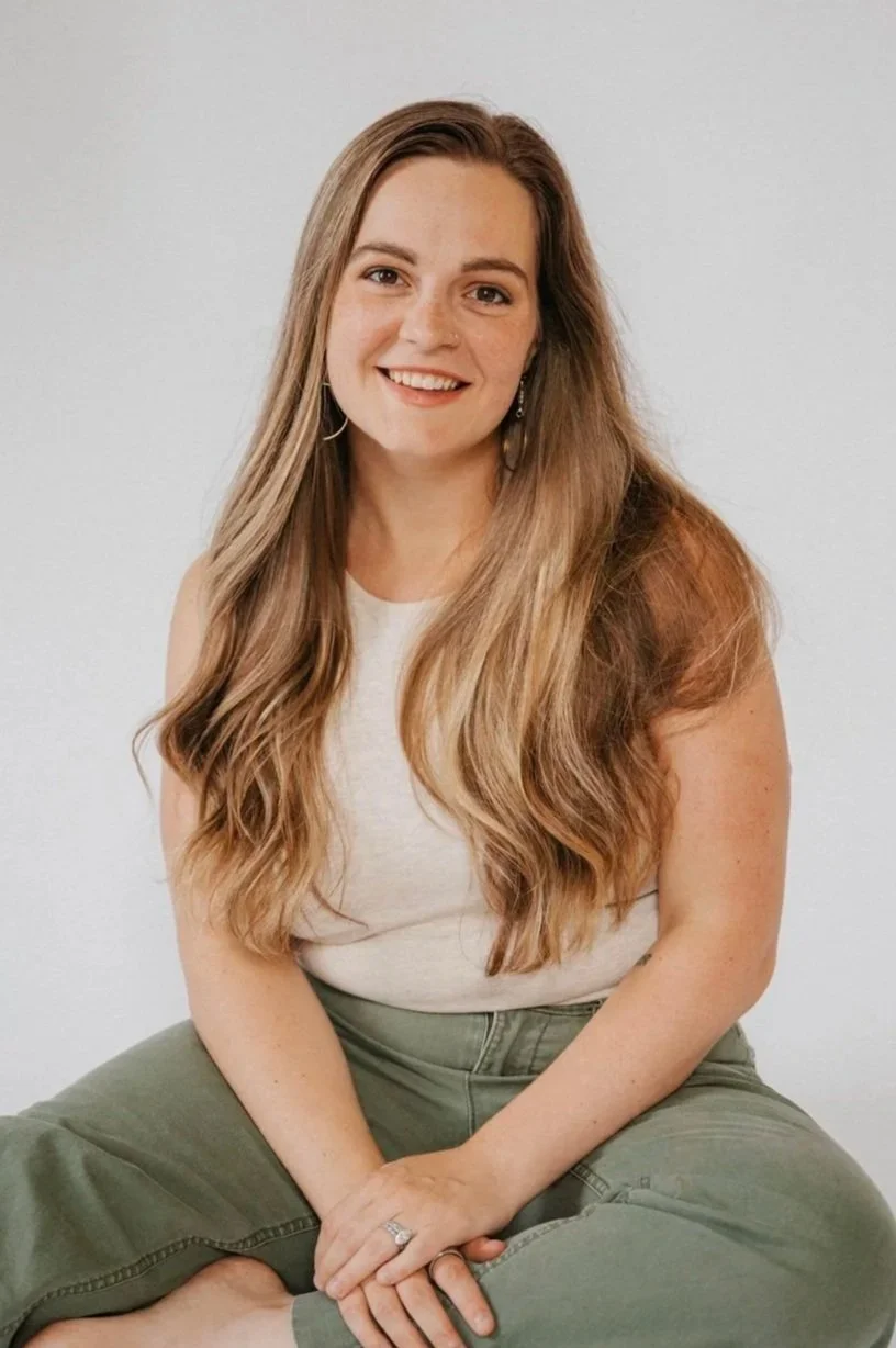A young woman with long, wavy, light brown hair, sitting cross-legged against a plain white background, smiling at the camera, wearing a white sleeveless top and olive green pants.
