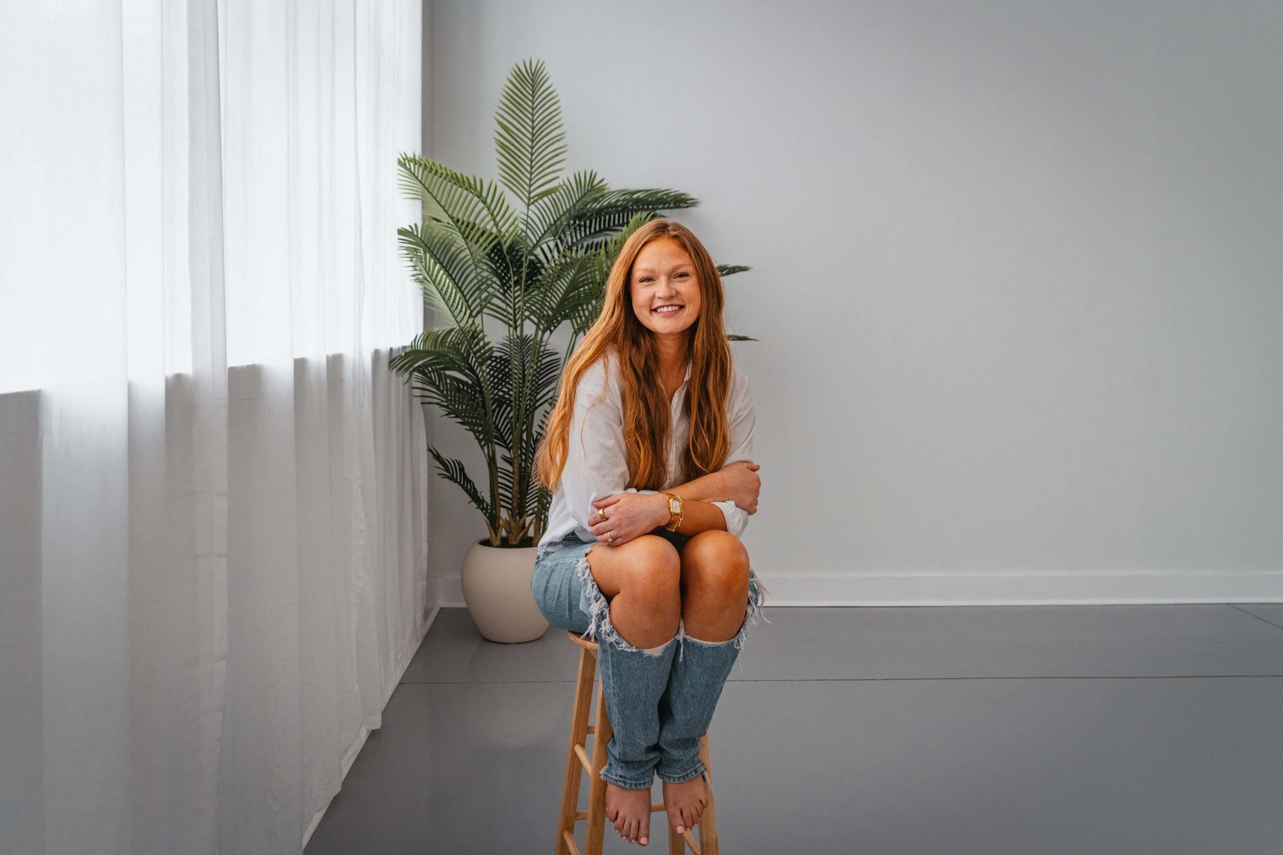 A young woman with long red hair, smiling, sitting on a wooden stool with her legs crossed, in a bright room with white walls, a large potted plant behind her, and sheer white curtains by the window.