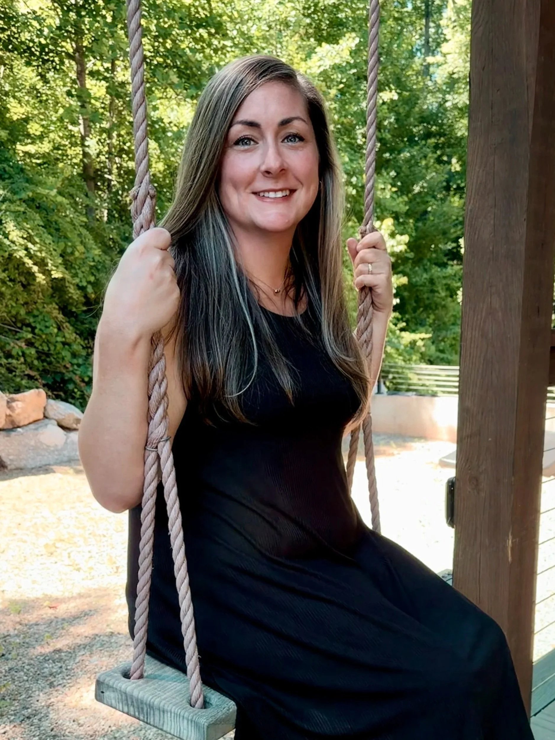 A smiling woman with long brown hair sitting on a swing with ropes, in an outdoor setting with trees and greenery in the background.