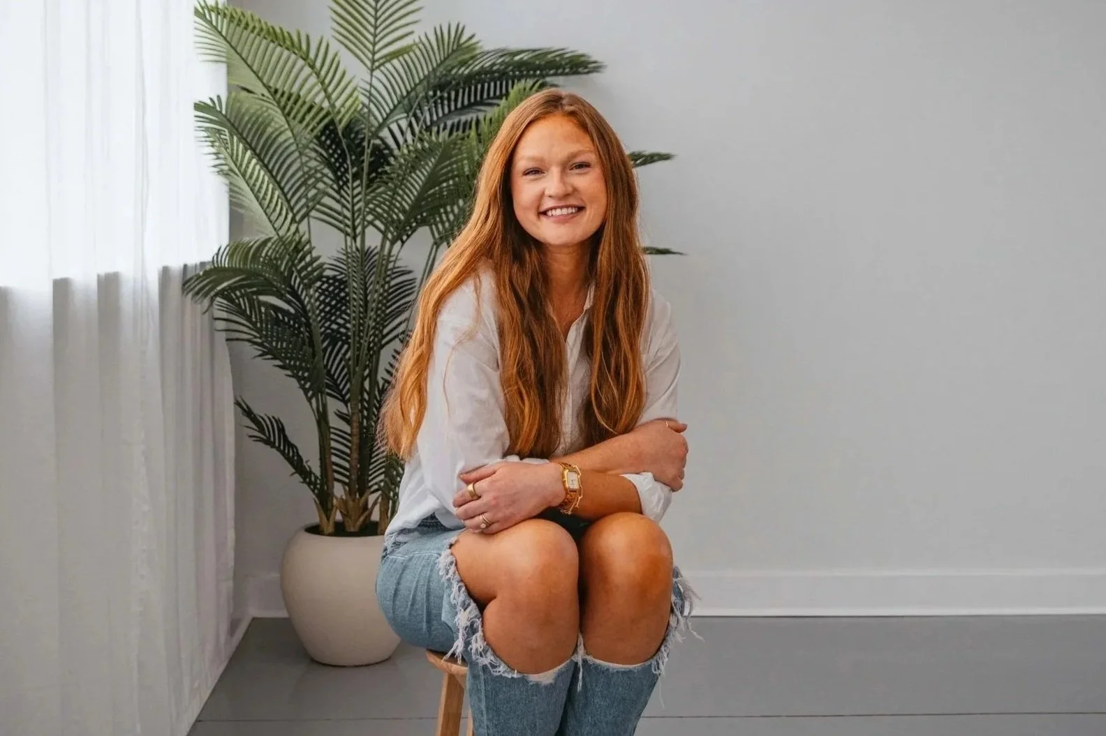 A young woman with long red hair, wearing a white long-sleeve shirt and ripped denim shorts, sitting on a stool with her arms crossed, smiling in front of a large green potted plant and a gray wall.