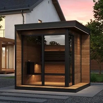 A small modern outdoor sauna with a black metal frame and wooden exterior, featuring large glass sliding doors, situated on a concrete pad in a backyard with a house and trees in the background.
