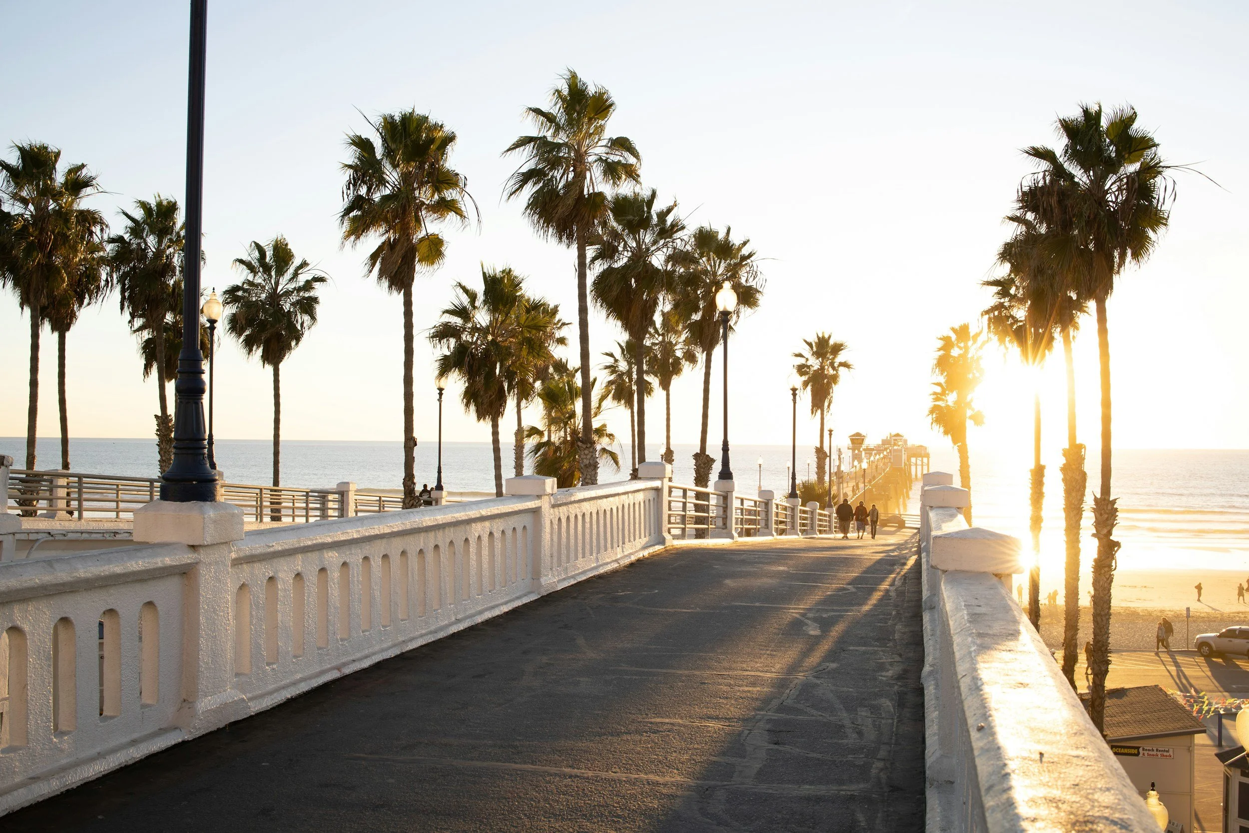 Sunset at a pier with palm trees, a white railing, and a few people walking along the boardwalk toward the ocean.