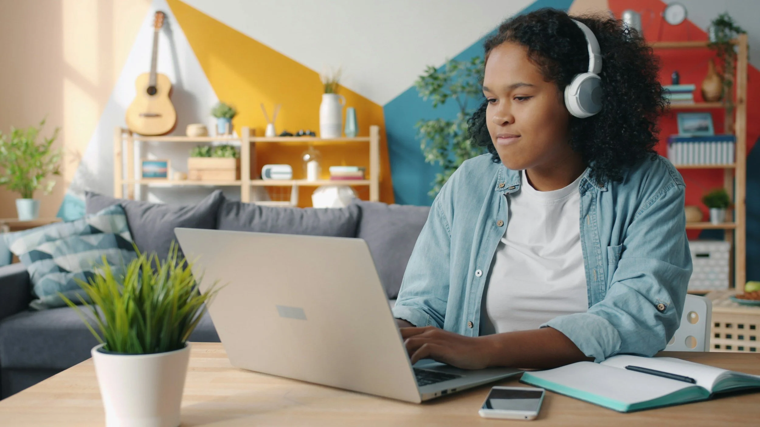 A young woman working on a laptop at a desk with a notebook, pen, and smartphone, wearing headphones in a colorful living room.