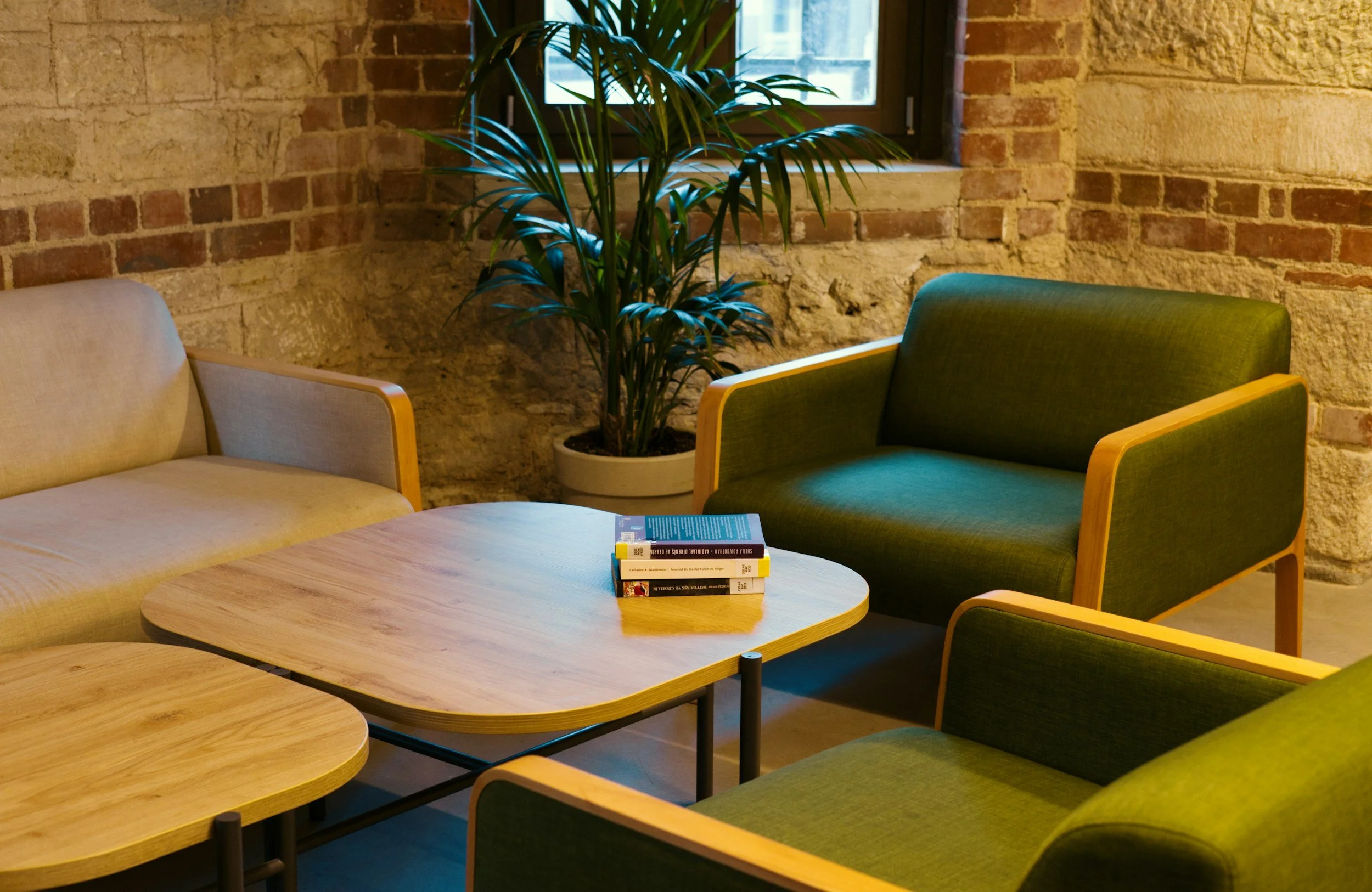 A cozy sitting area with a beige sofa and green armchair arranged around a light wood coffee table. A potted plant with large green leaves is placed near the window behind the furniture. The background features exposed brick walls and a window, creating an inviting atmosphere.