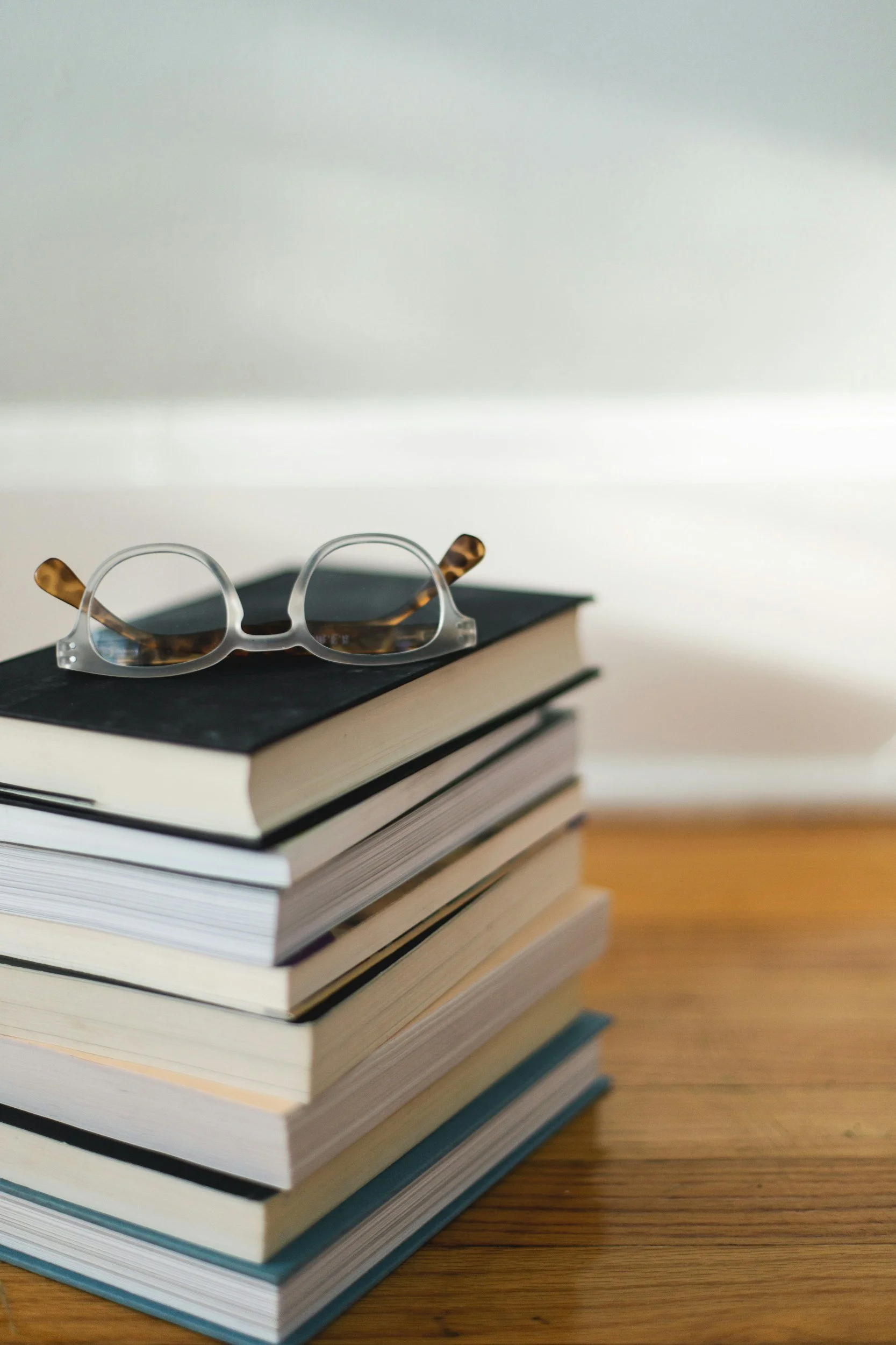 A stack of books with a pair of eyeglasses on top, placed on a wooden surface.