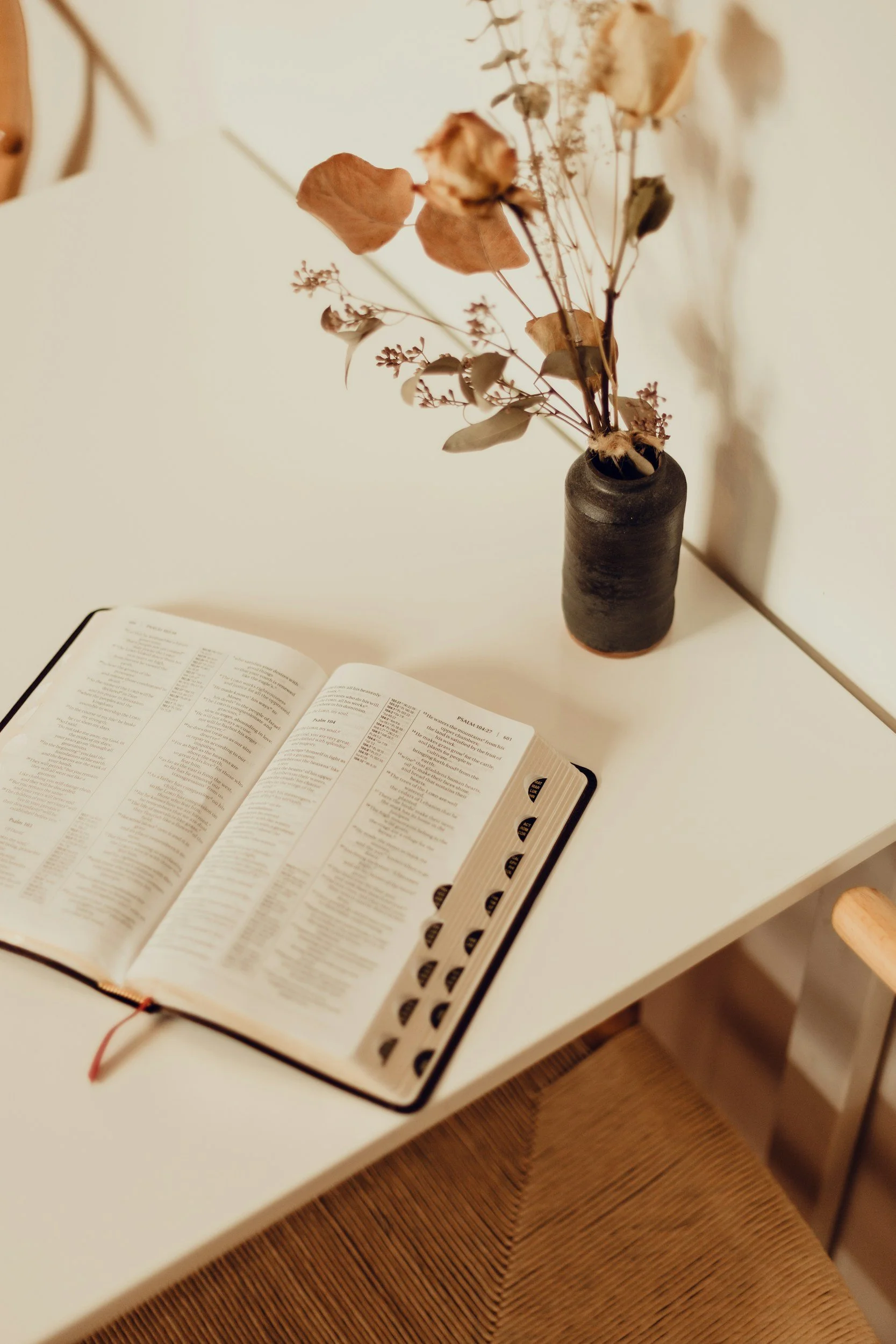 An open Bible on a white table with a black vase holding dried flowers nearby.