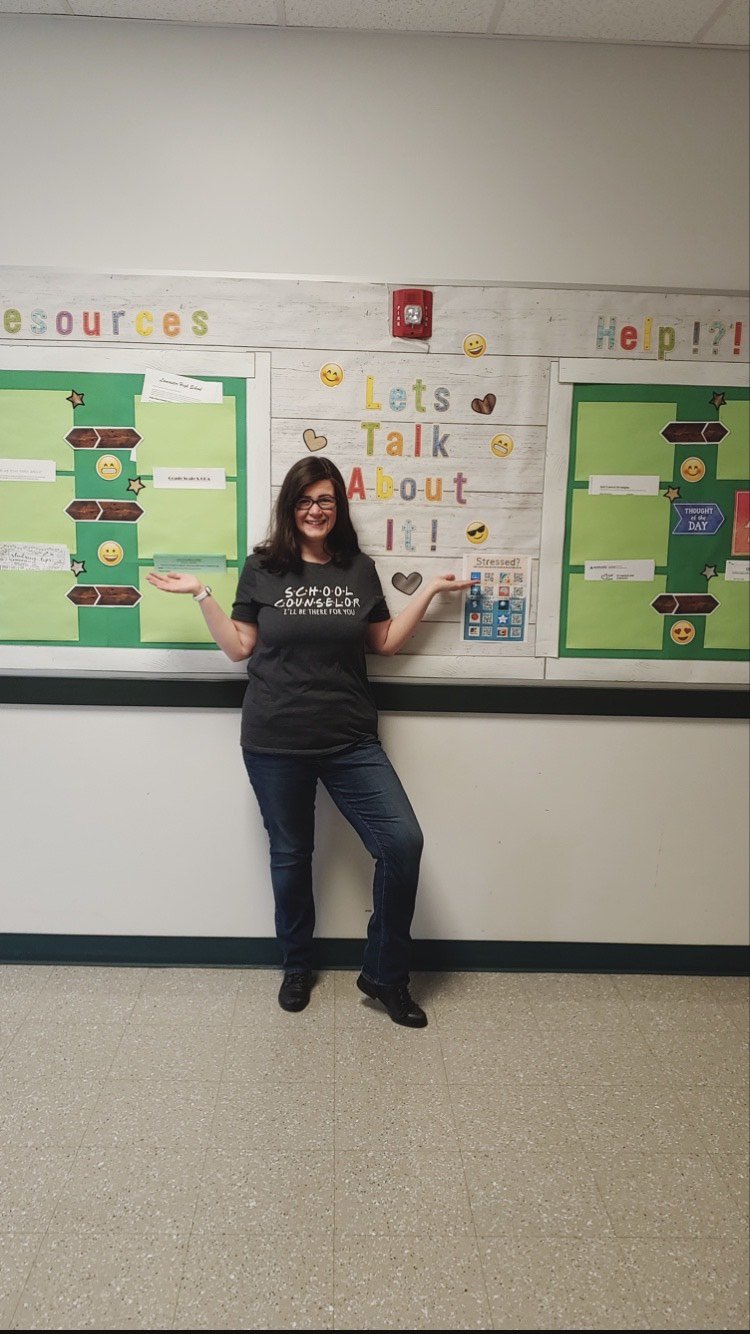 Woman standing in front of a school bulletin board that reads 'Let's Talk About It!' with colorful letters and emojis, smiling and posing with arms outstretched.
