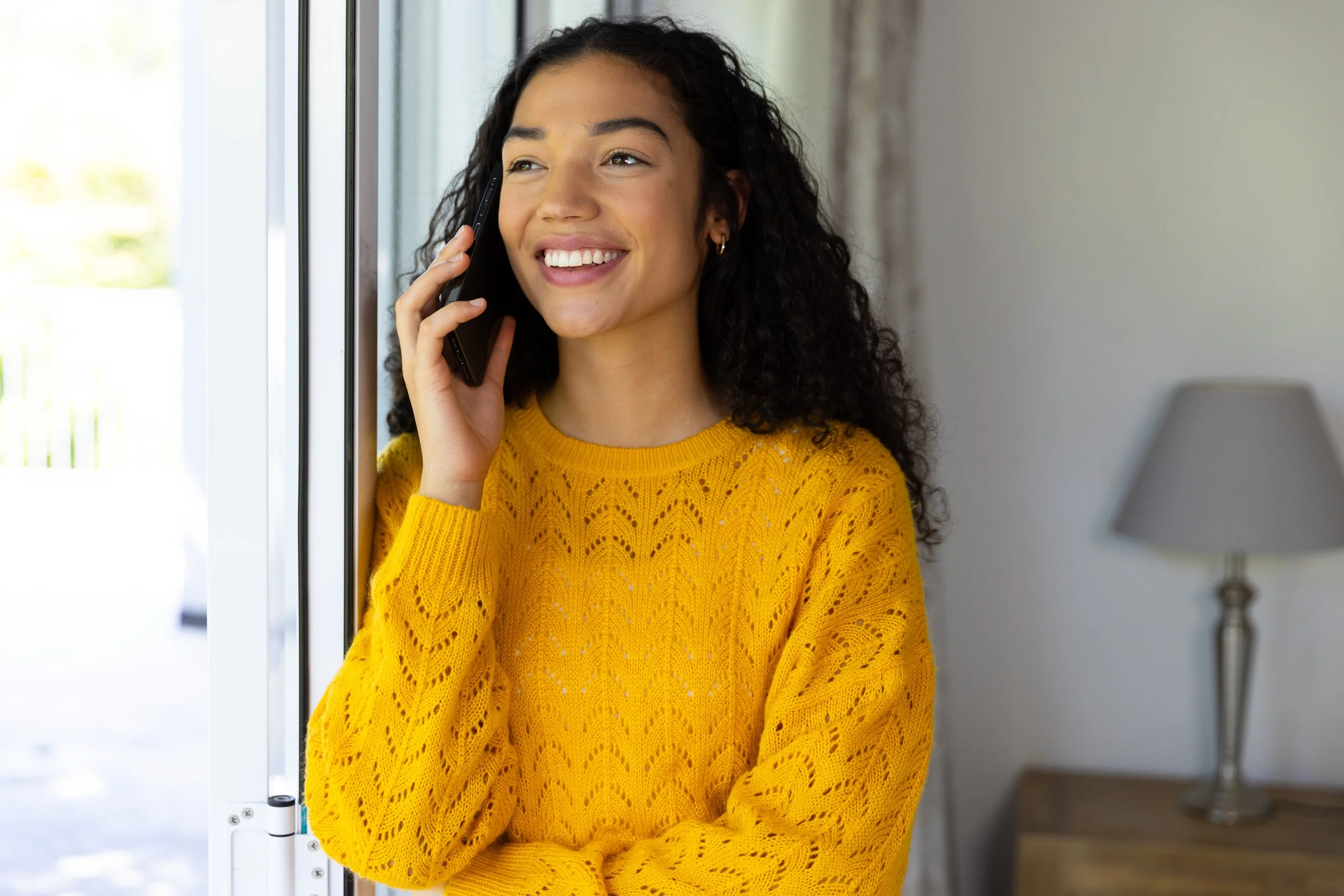 A young woman with curly black hair wearing a yellow sweater, talking on a cellphone indoors near a window, smiling.