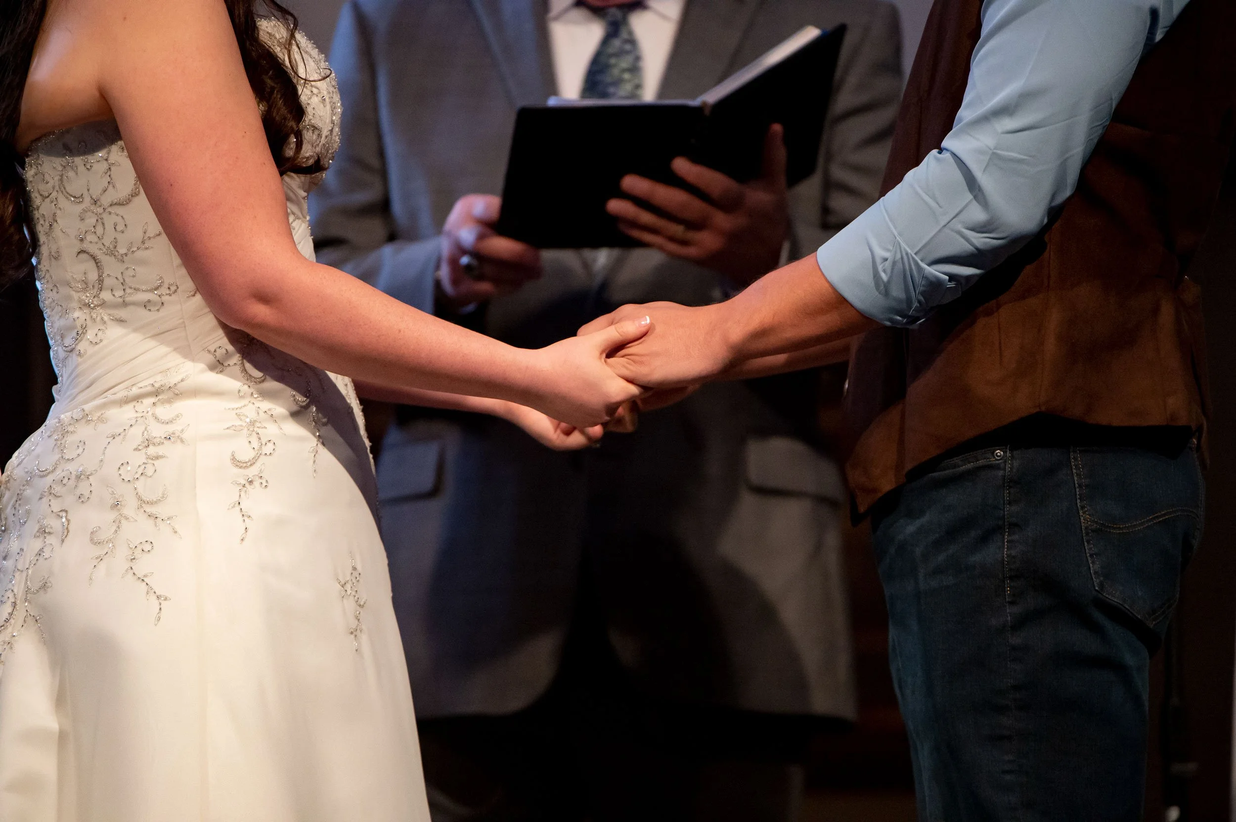 A bride and groom holding hands during their wedding ceremony, with an officiant reading from a book in the background.