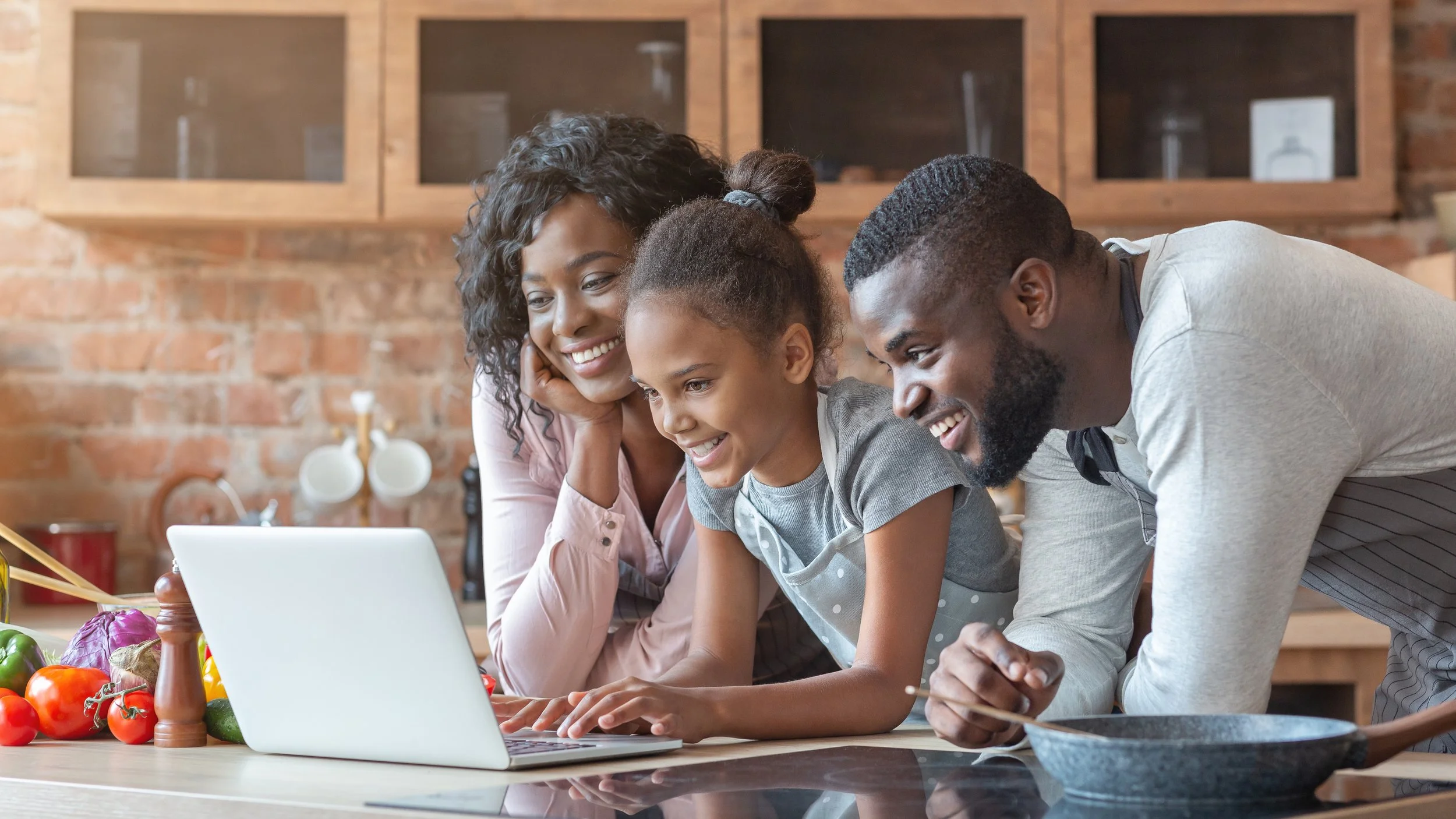 A family looking at a laptop in the kitchen with fresh vegetables on the counter.