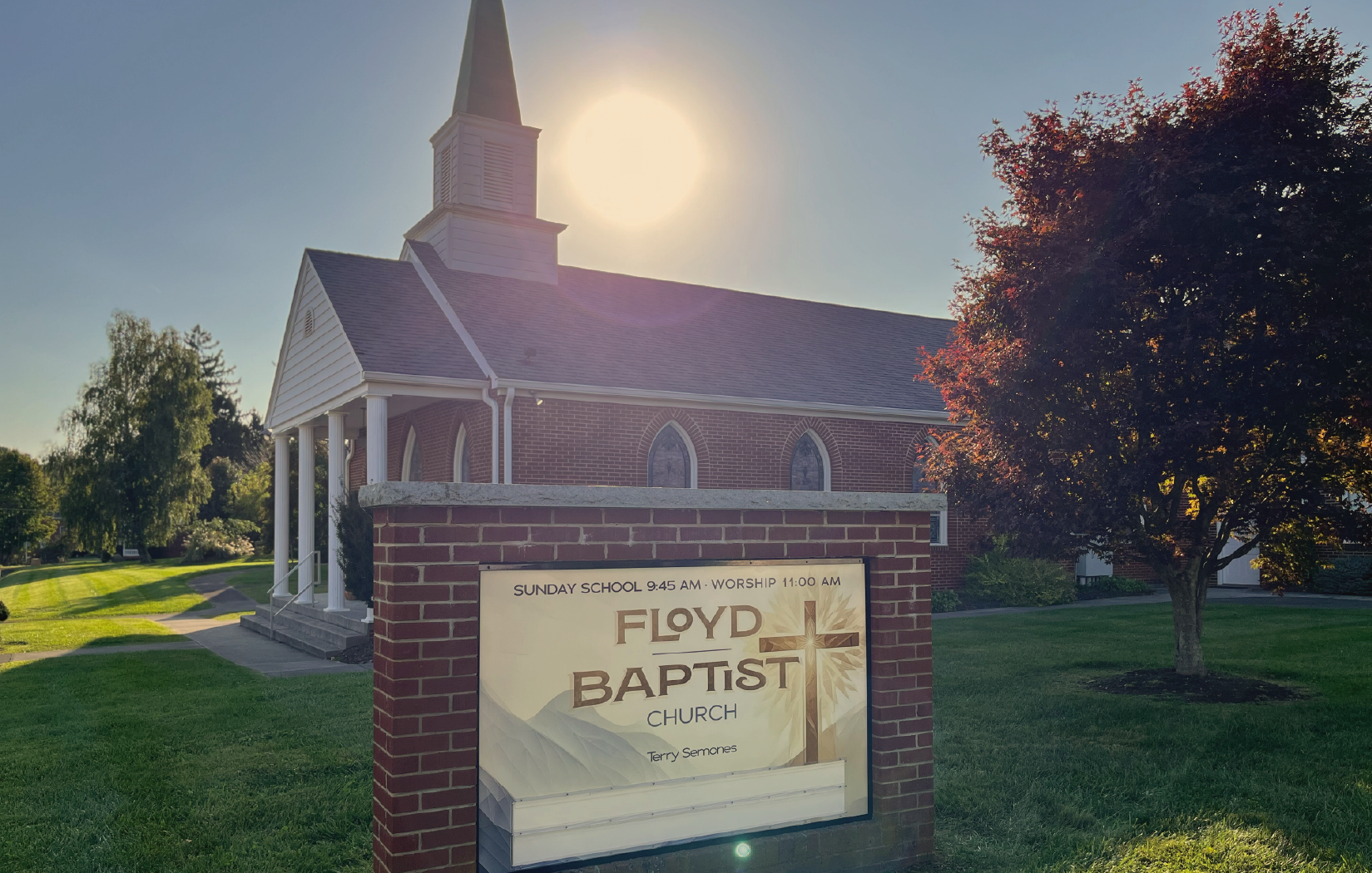 A brick church with a steeple and a sign in front that reads 'Floyd Baptist Church' with service times, under a bright sun with a clear sky and trees in the background.