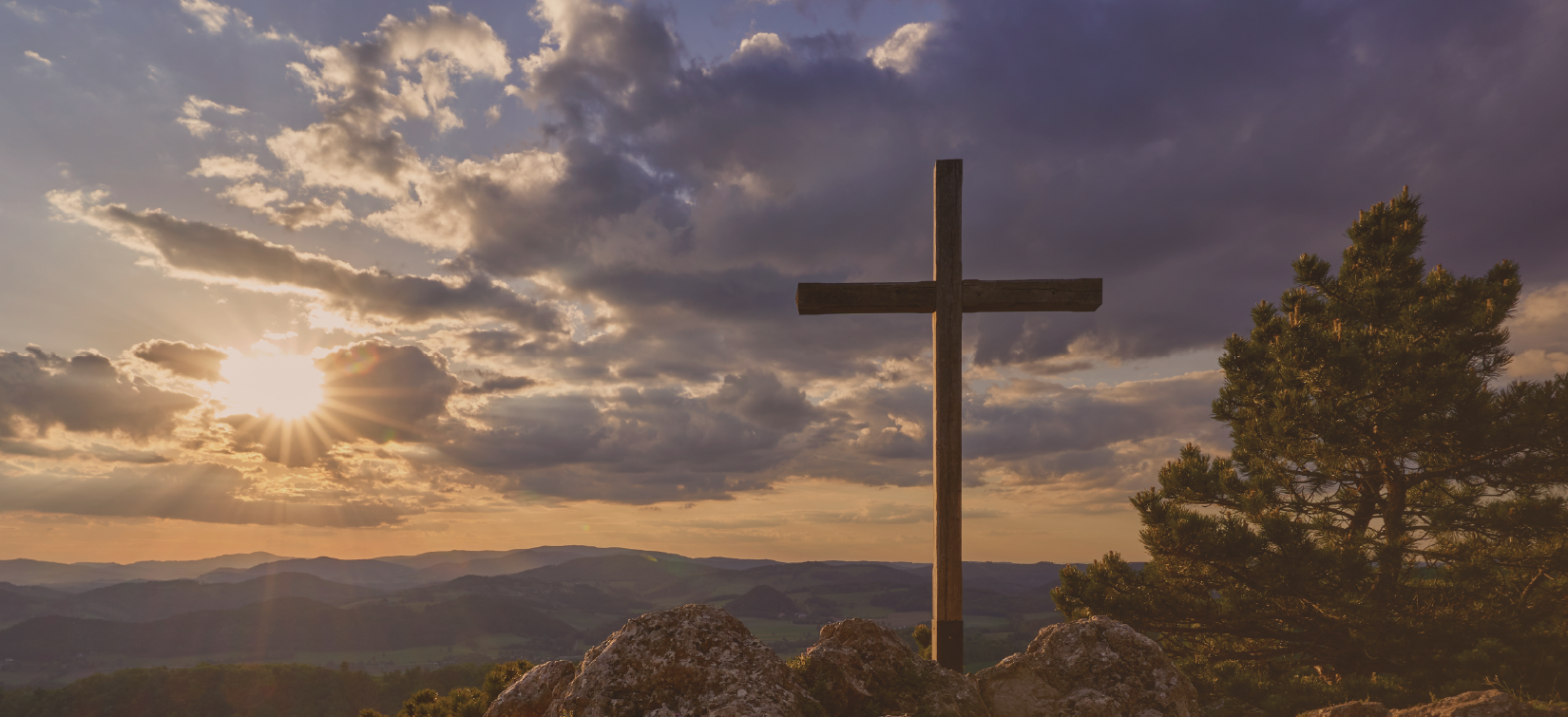 A wooden cross on a rocky hilltop during sunset, with a pine tree on the right, cloudy sky, and mountains in the background.