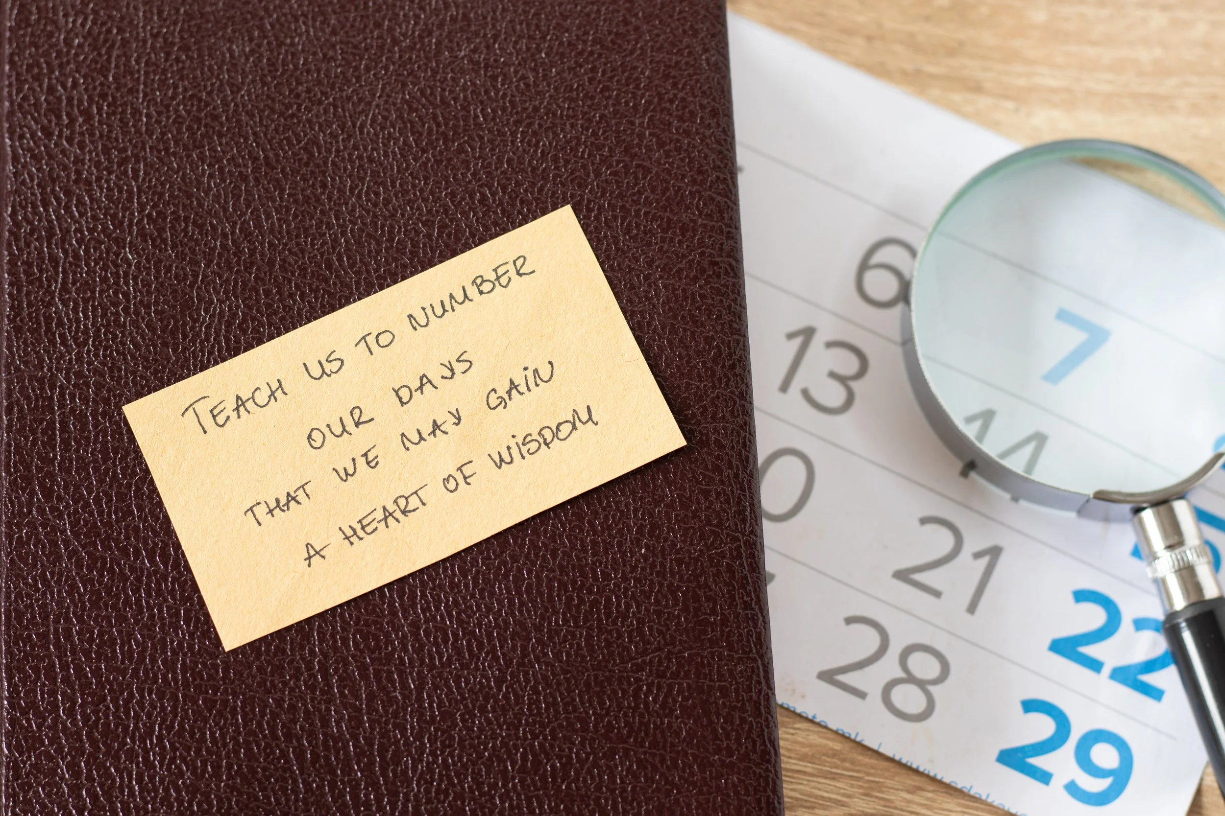 A yellow sticky note with a handwritten quote is placed on a brown textured notebook. The quote reads, 'Teach us to number our days that we may gain a heart of wisdom.' In the background, there is a calendar with dates and a magnifying glass partially over it.