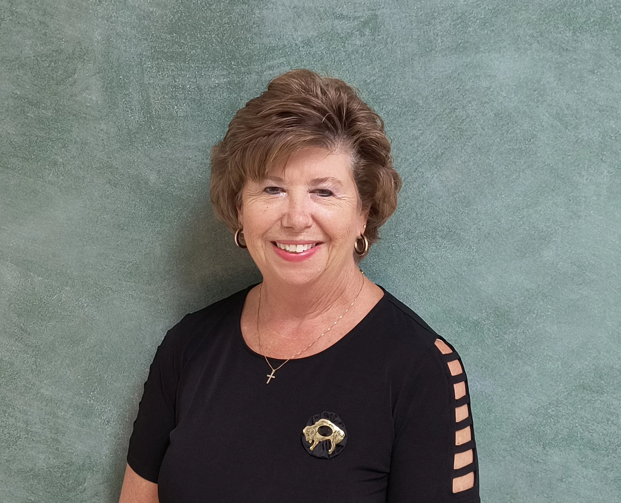A smiling middle-aged woman with short, wavy brown hair, wearing a black top with cut-out shoulder details, gold earrings, a gold cross necklace, and a decorative brooch on her left chest, standing against a green wall.