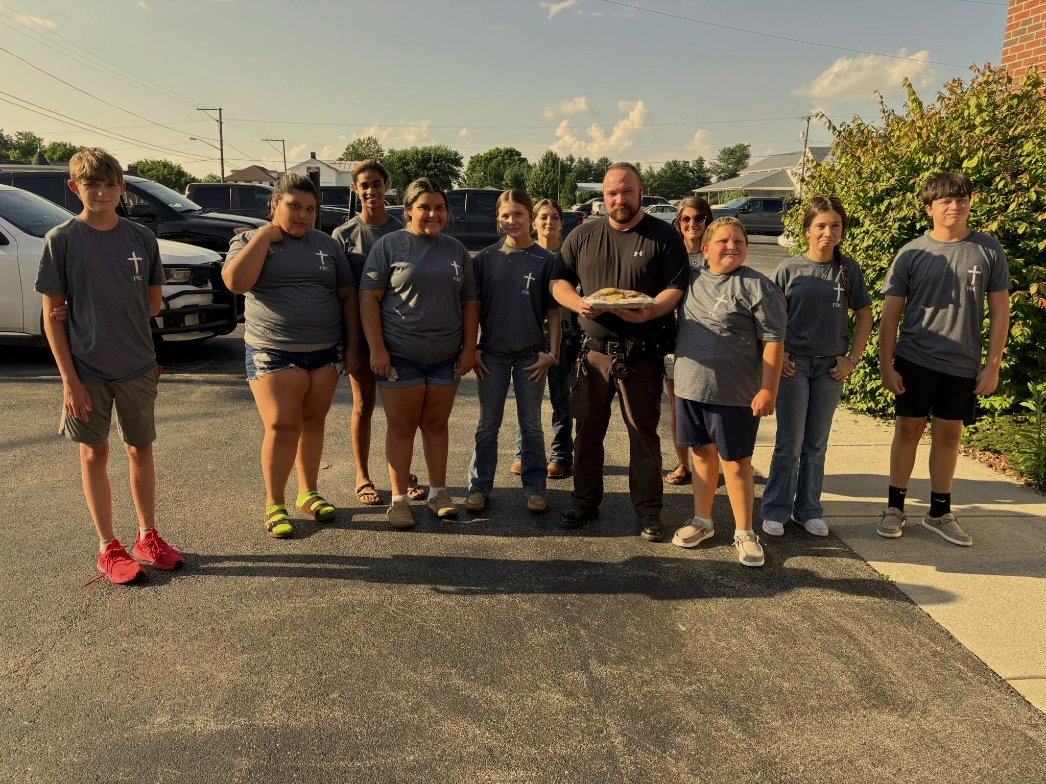A group of ten people standing outdoors in a parking lot, with a man holding a tray of bread in the center. The group includes young people and adults, some wearing matching gray shirts with a cross logo.