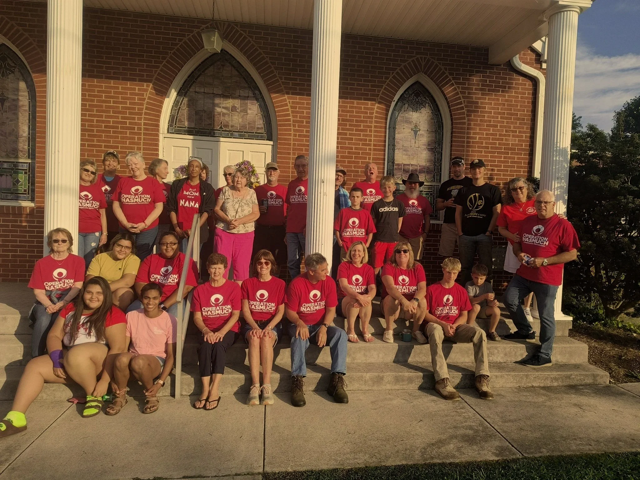 Group of people gathered on the steps of a brick building with church-like architecture, some wearing red T-shirts with 'Operation NASMUCH' printed on them, posing for a group photo.