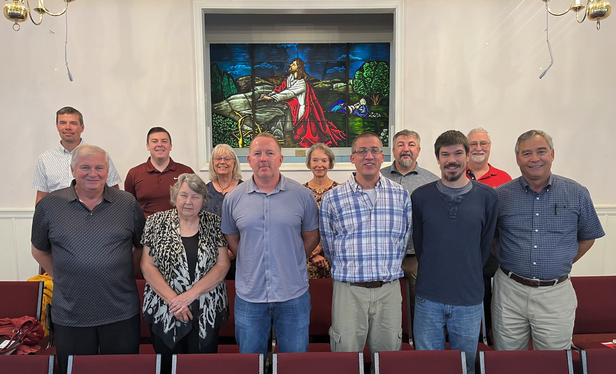 A group of eleven people standing and sitting in a room, with a stained glass window depicting Jesus in the background.