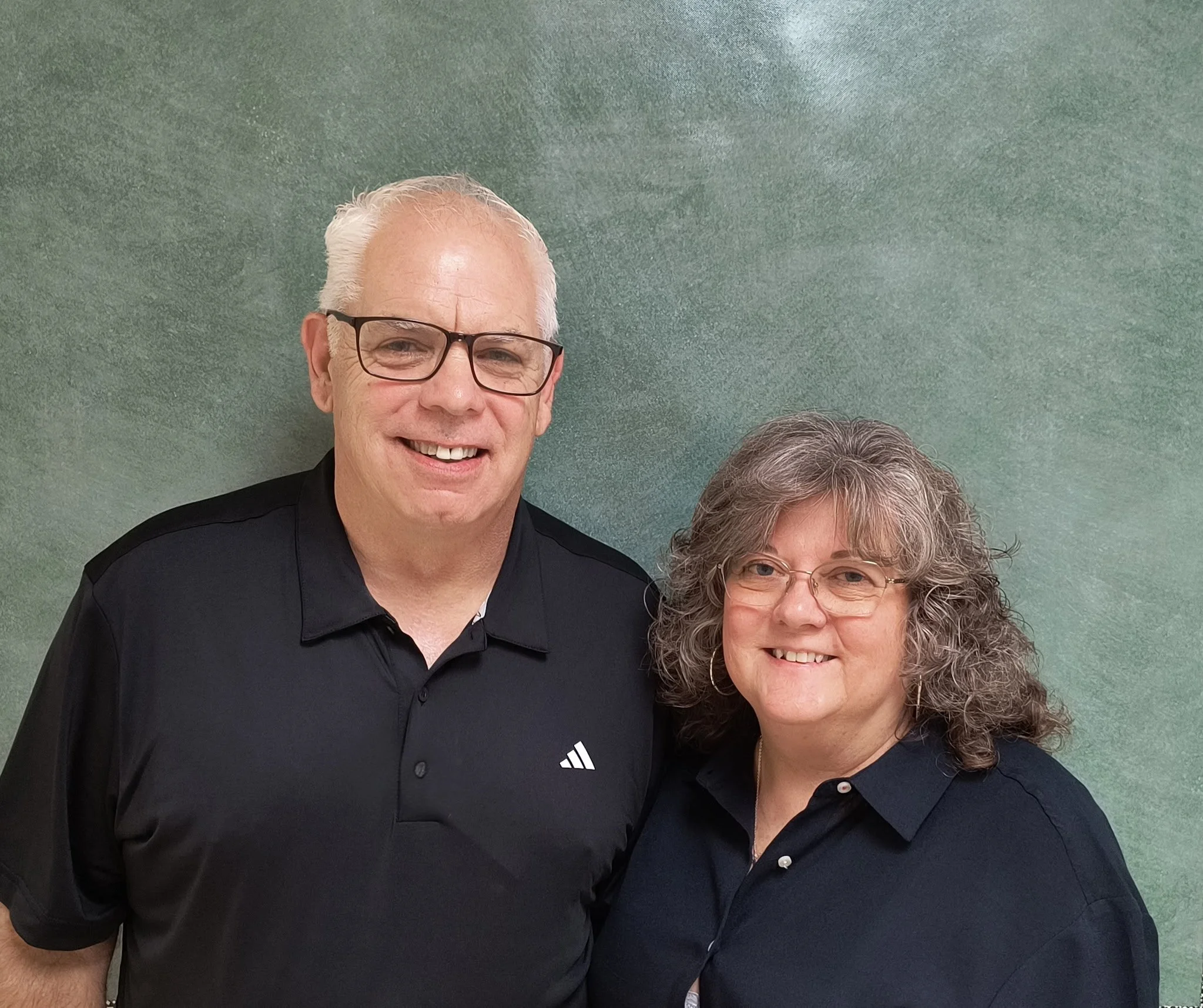 A smiling man with glasses and a woman with curly hair and glasses standing together against a green background.