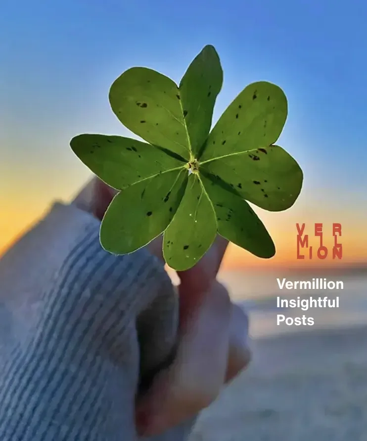A close up photo of a person's hand holding a rare four-leaf clover with heart shaped leaves with a sunset and beach in the background.