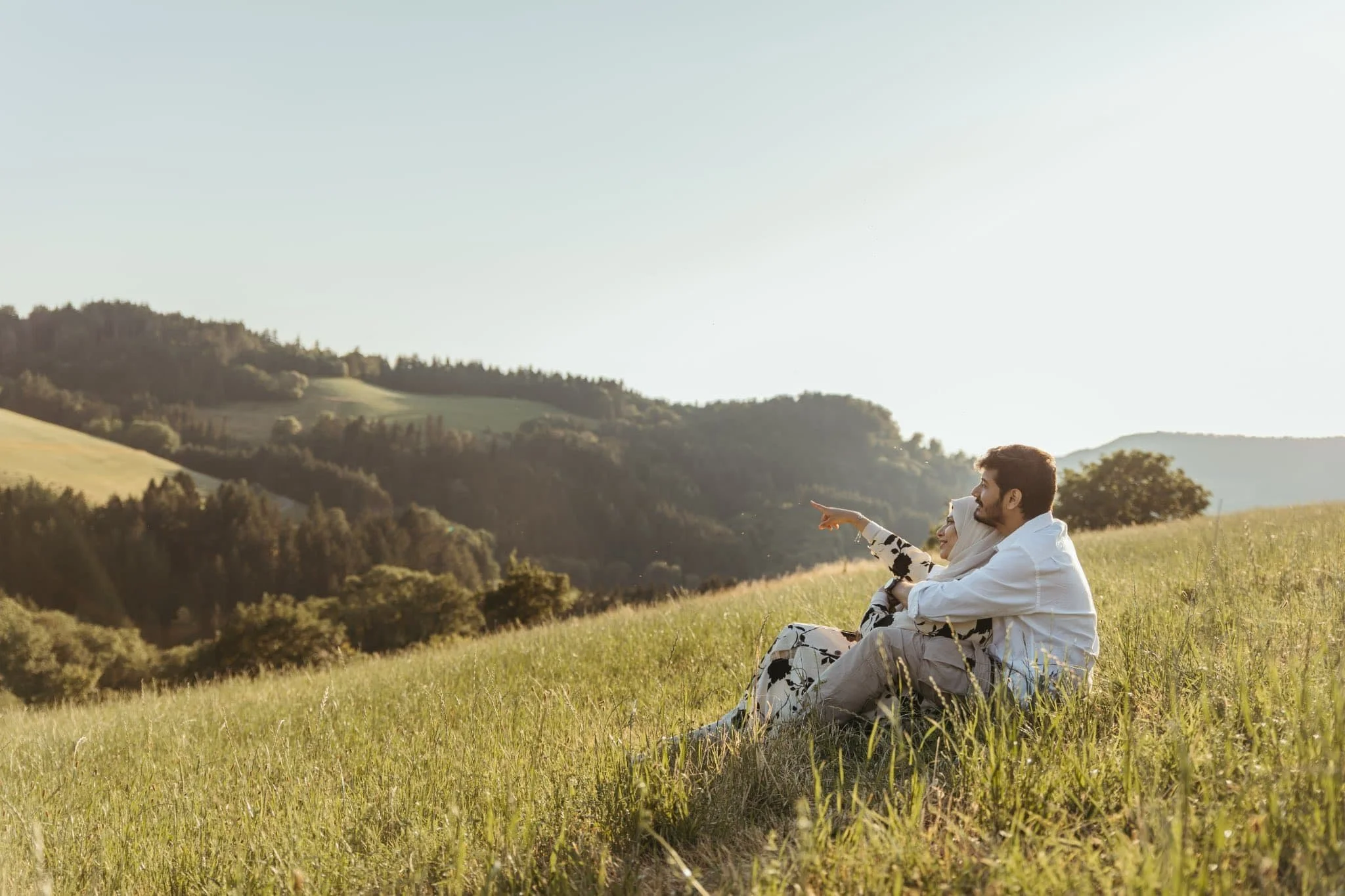 A couple sitting on a grassy hill, with the woman pointing towards the distant landscape, in a scenic countryside during sunset in connecticut