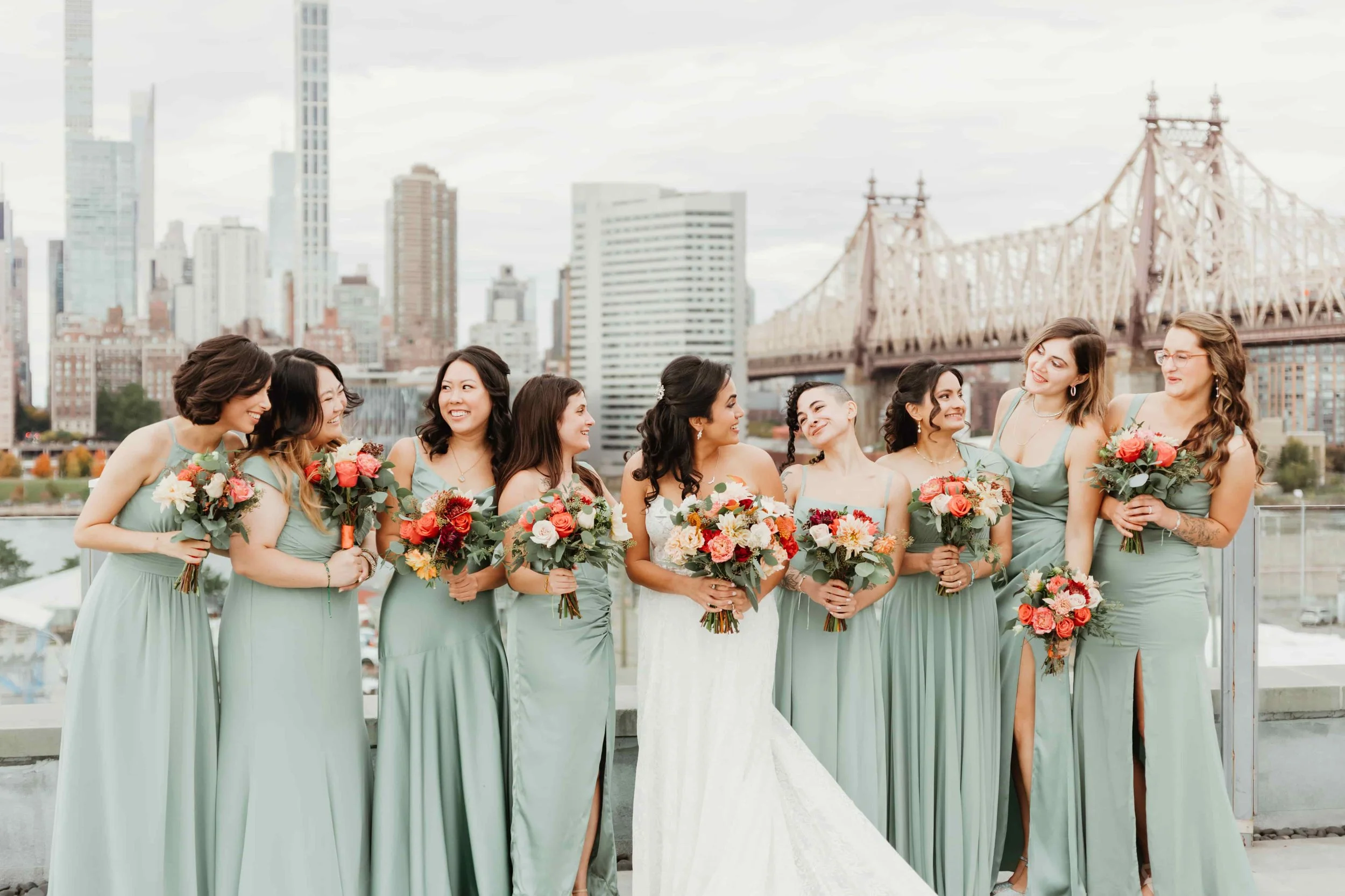 Bride and bridesmaids in matching green dresses holding bouquets, standing on a rooftop in New York City with a bridge and skyscrapers in the NYC
