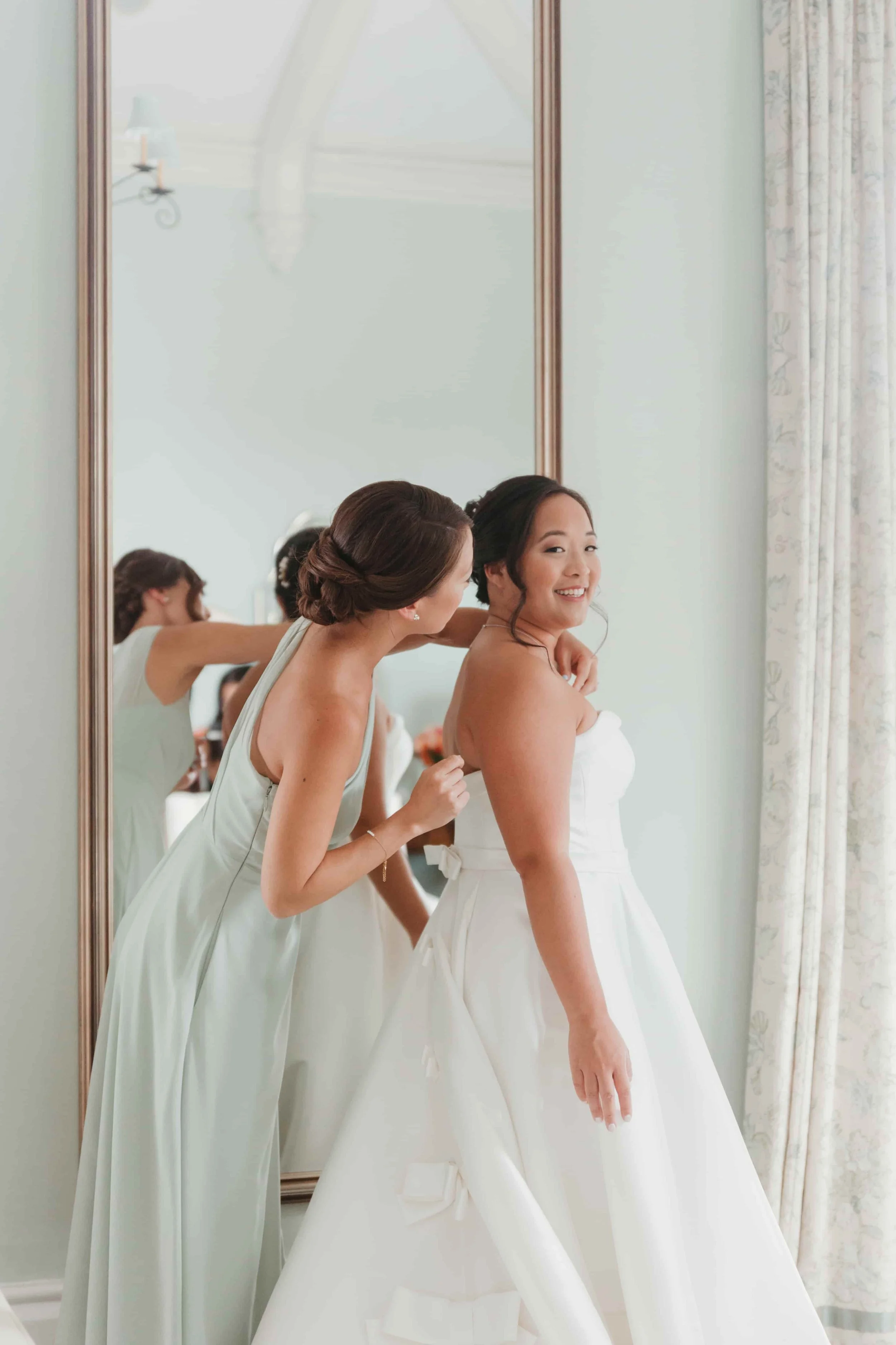A bride in a white wedding gown smiling as her friend helps her prepare, reflected in a large mirror in a well-lit room in connecticut