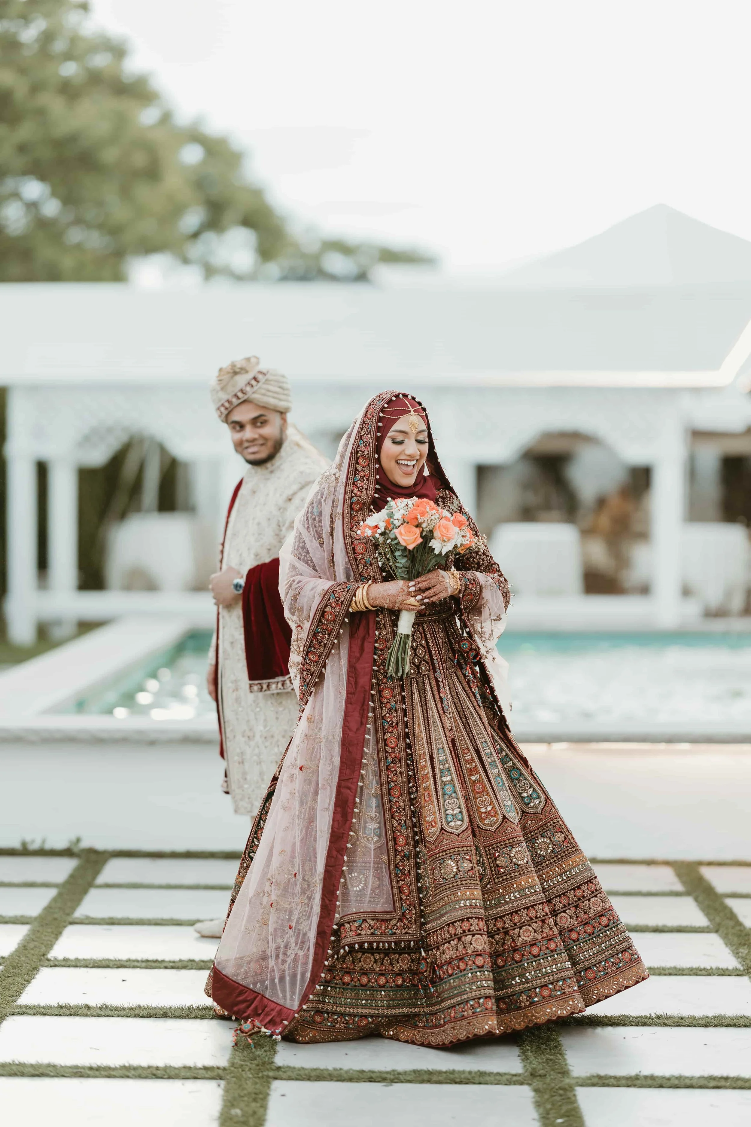 A woman dressed in traditional Indian wedding attire holding a bouquet of flowers, smiling, with a man in traditional attire standing behind her.