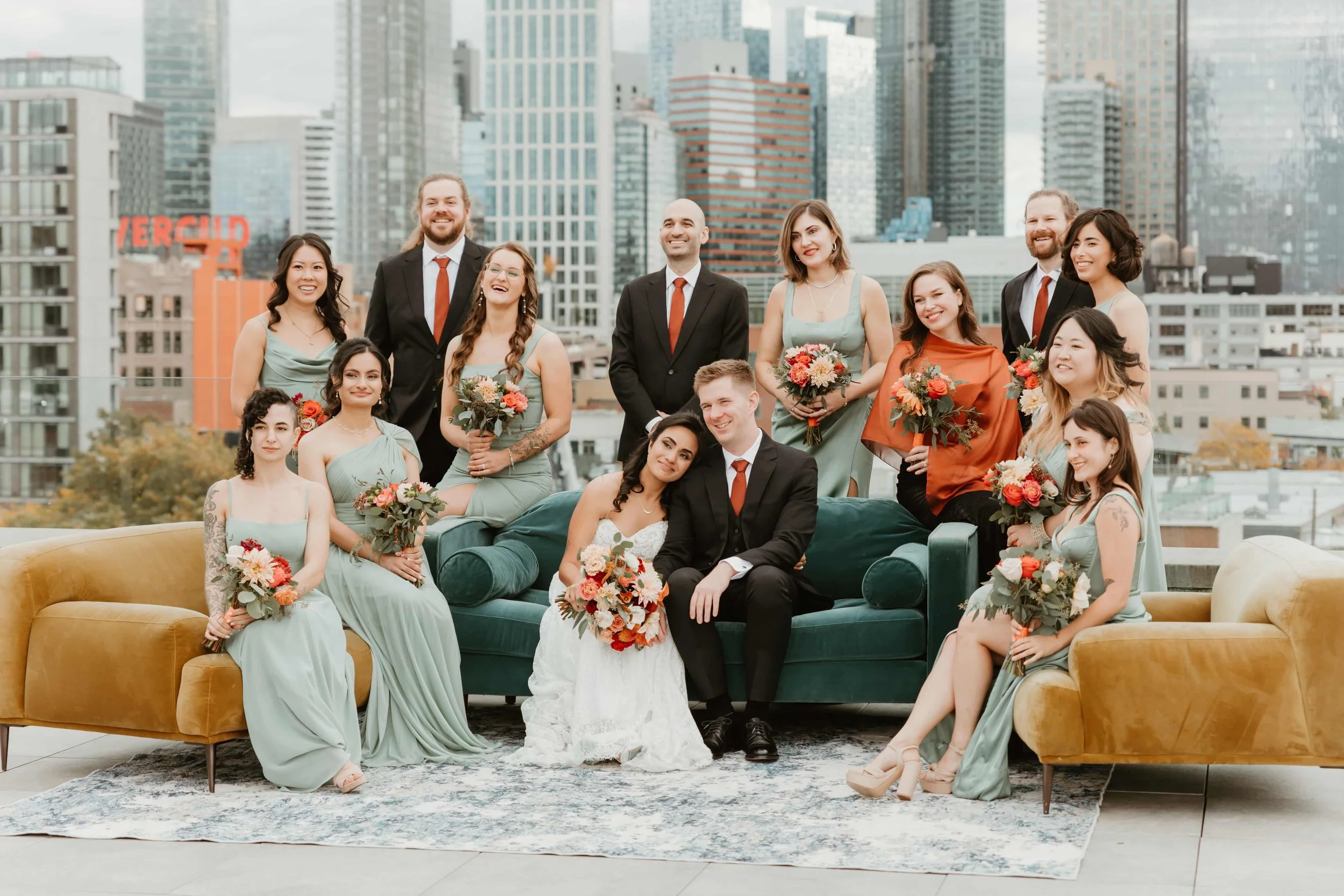 A wedding party with the bride and groom sitting on a green velvet sofa in front, surrounded by bridesmaids in mint green dresses, groomsmen in black suits with red ties, and other women holding bouquets, all on a rooftop with city skyscrapers in the background.