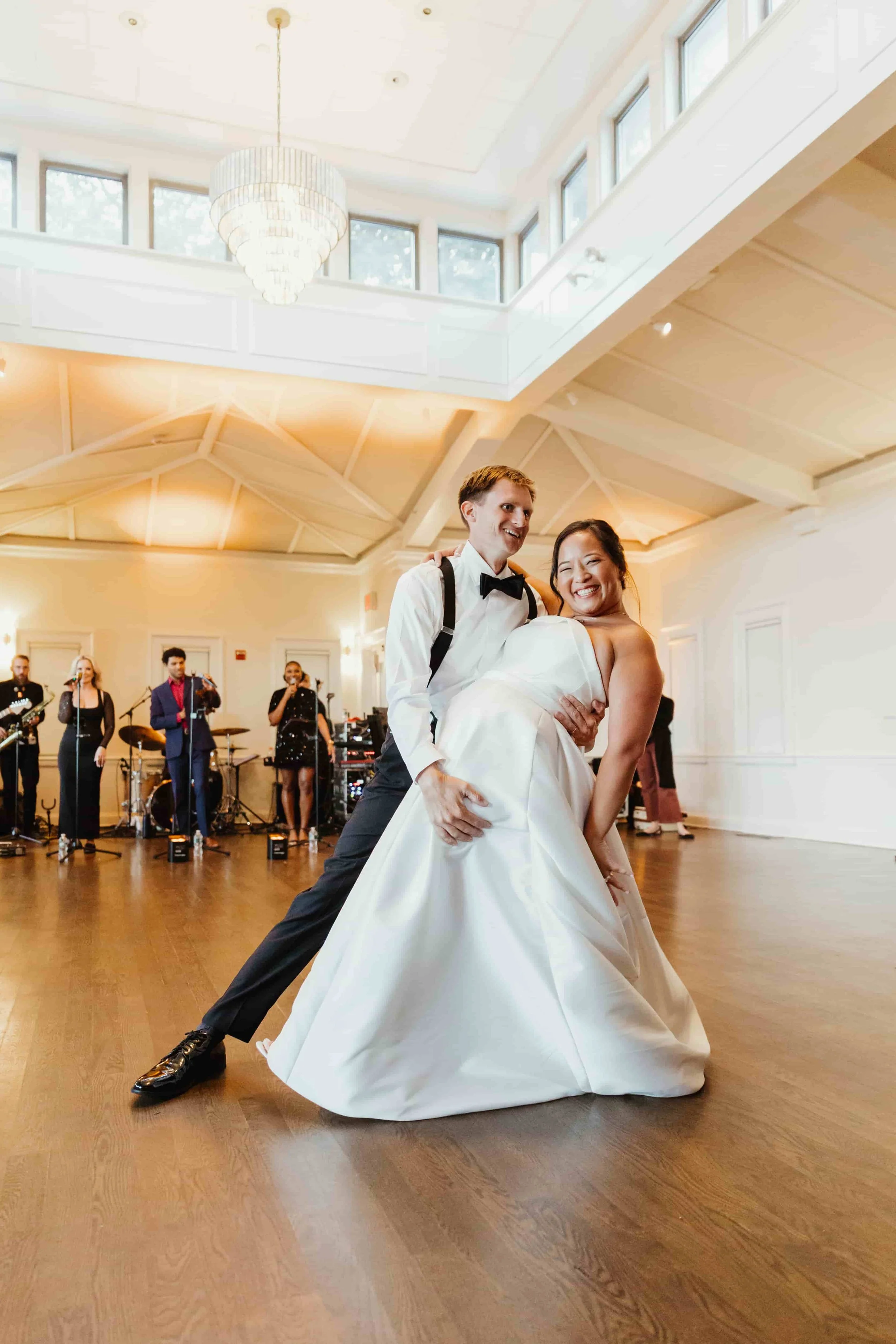 A bride and groom dancing at their wedding reception, with a band playing in the background inside a well-lit hall.