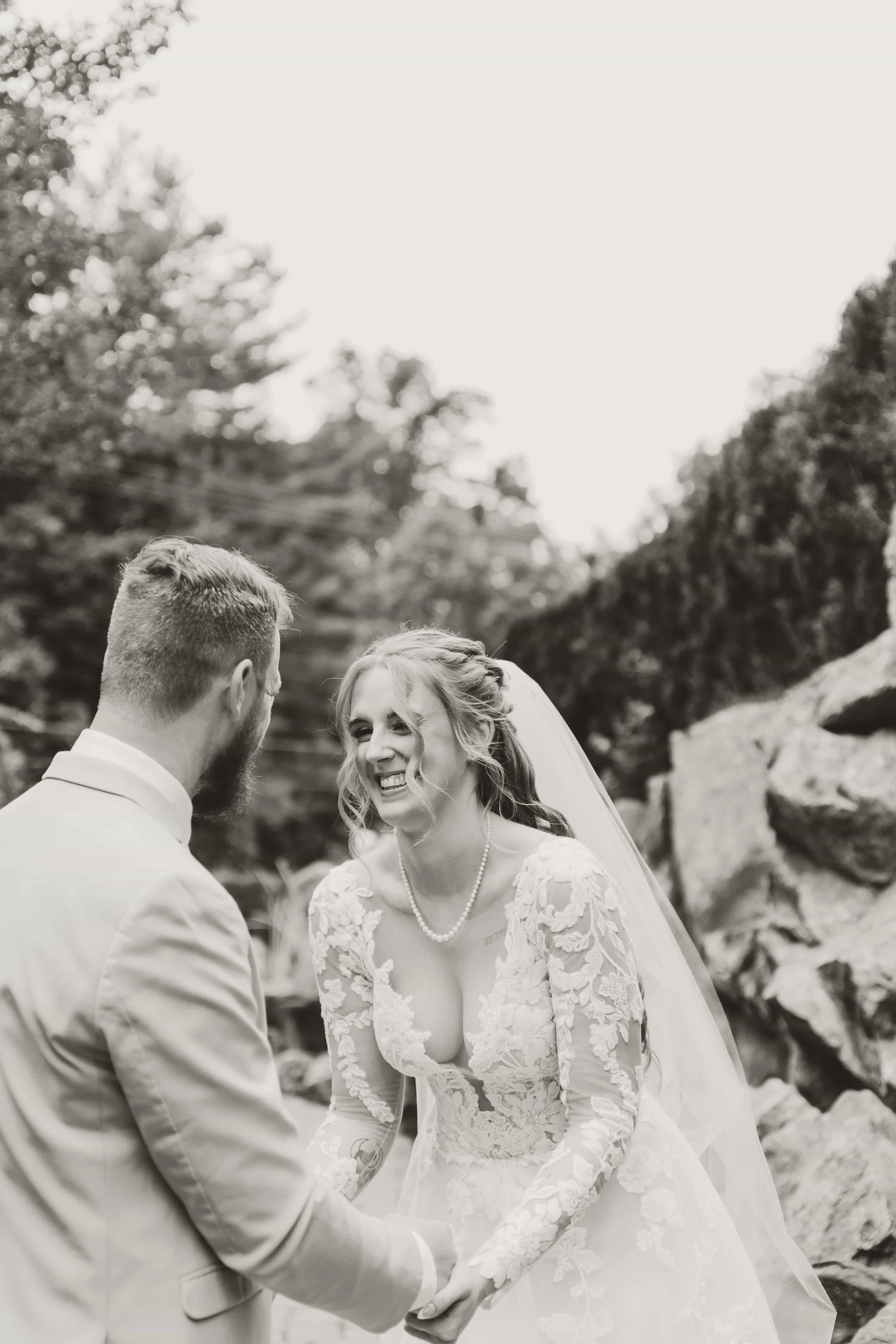 A black and white photo of a bride and groom holding hands outdoors, smiling and looking at each other.