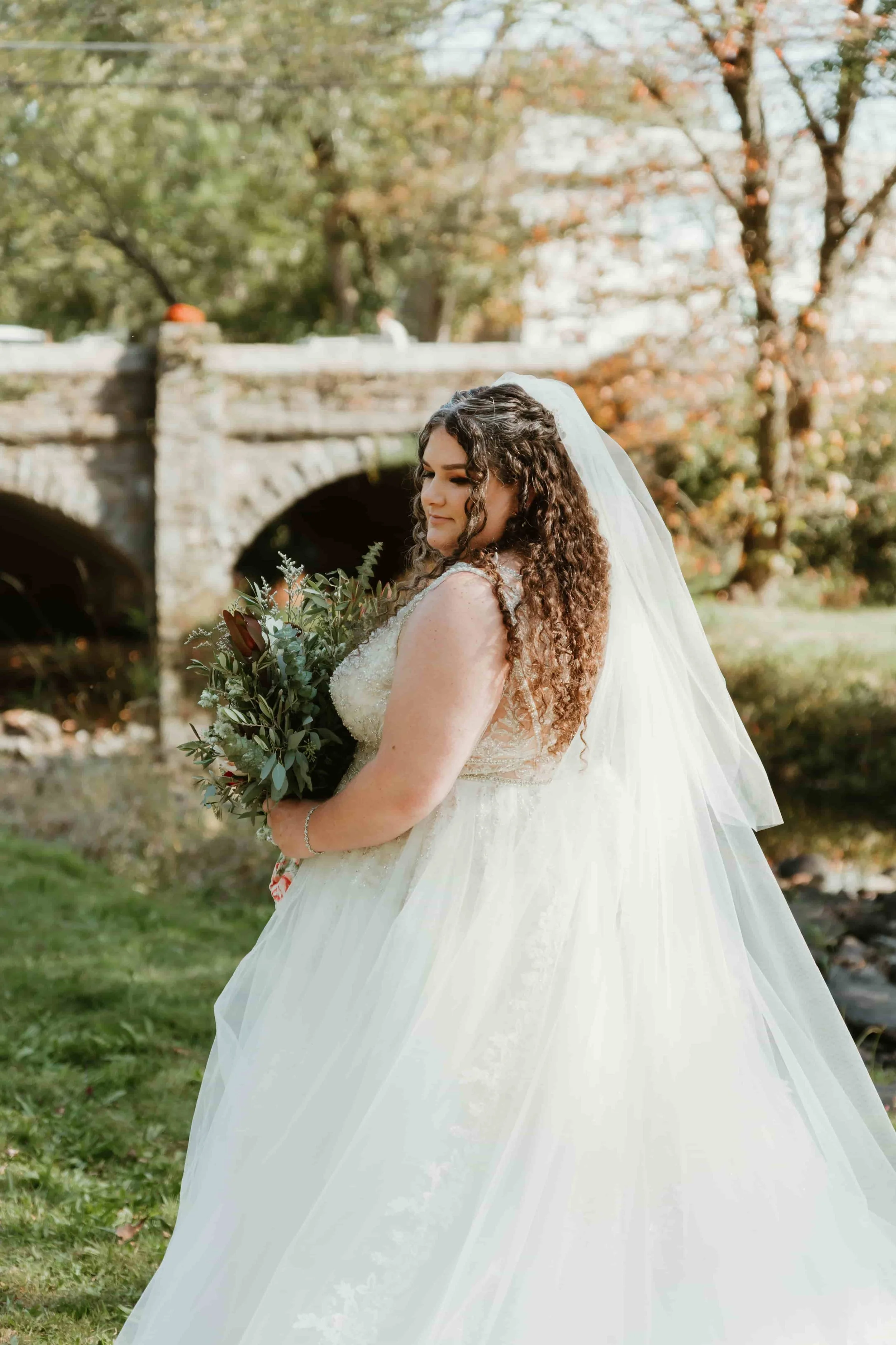 A bride with long curly hair wearing a white wedding dress and veil, standing outdoors in front of a stone bridge and autumn trees holding a bouquet of flowers.