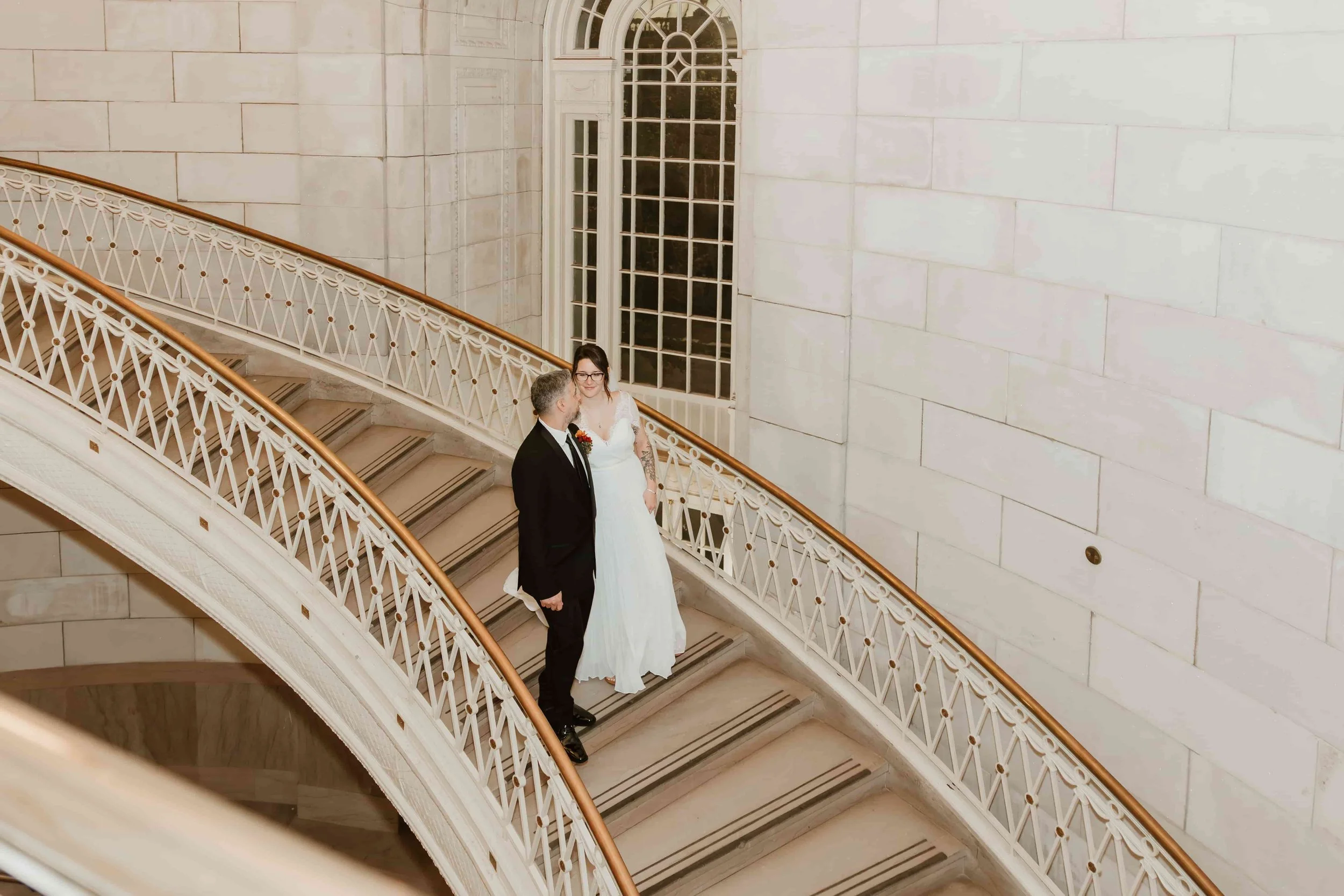 A bride and groom standing on a curved staircase with white railing and gold accents in front of large arched windows in a grand interior space.