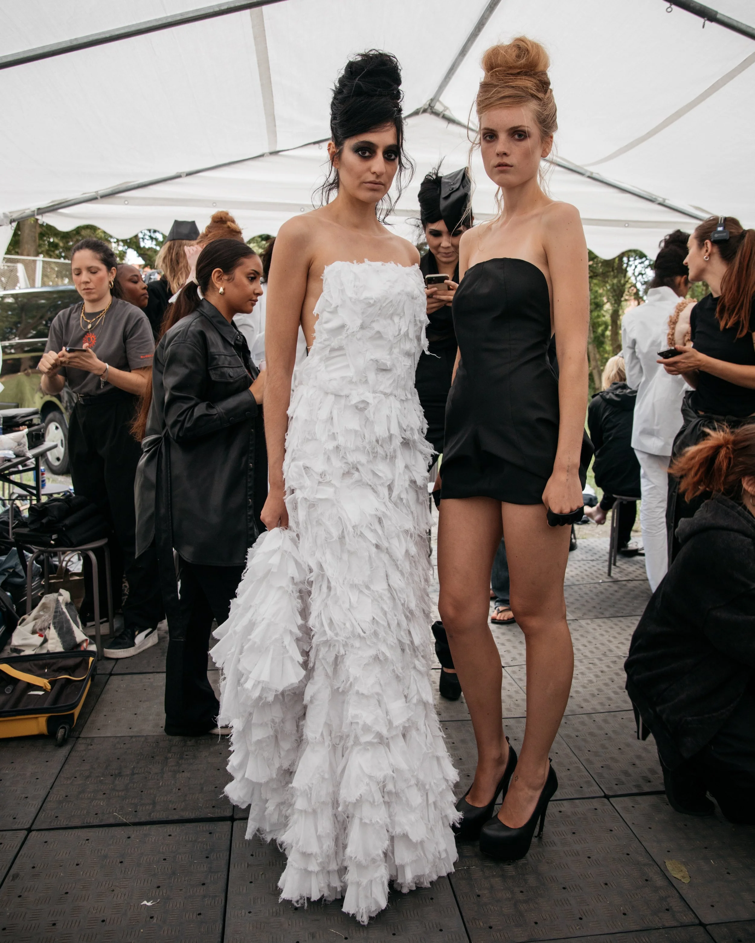 Two female models in designer outfits on a runway at a fashion show, surrounded by backstage staff and other models.